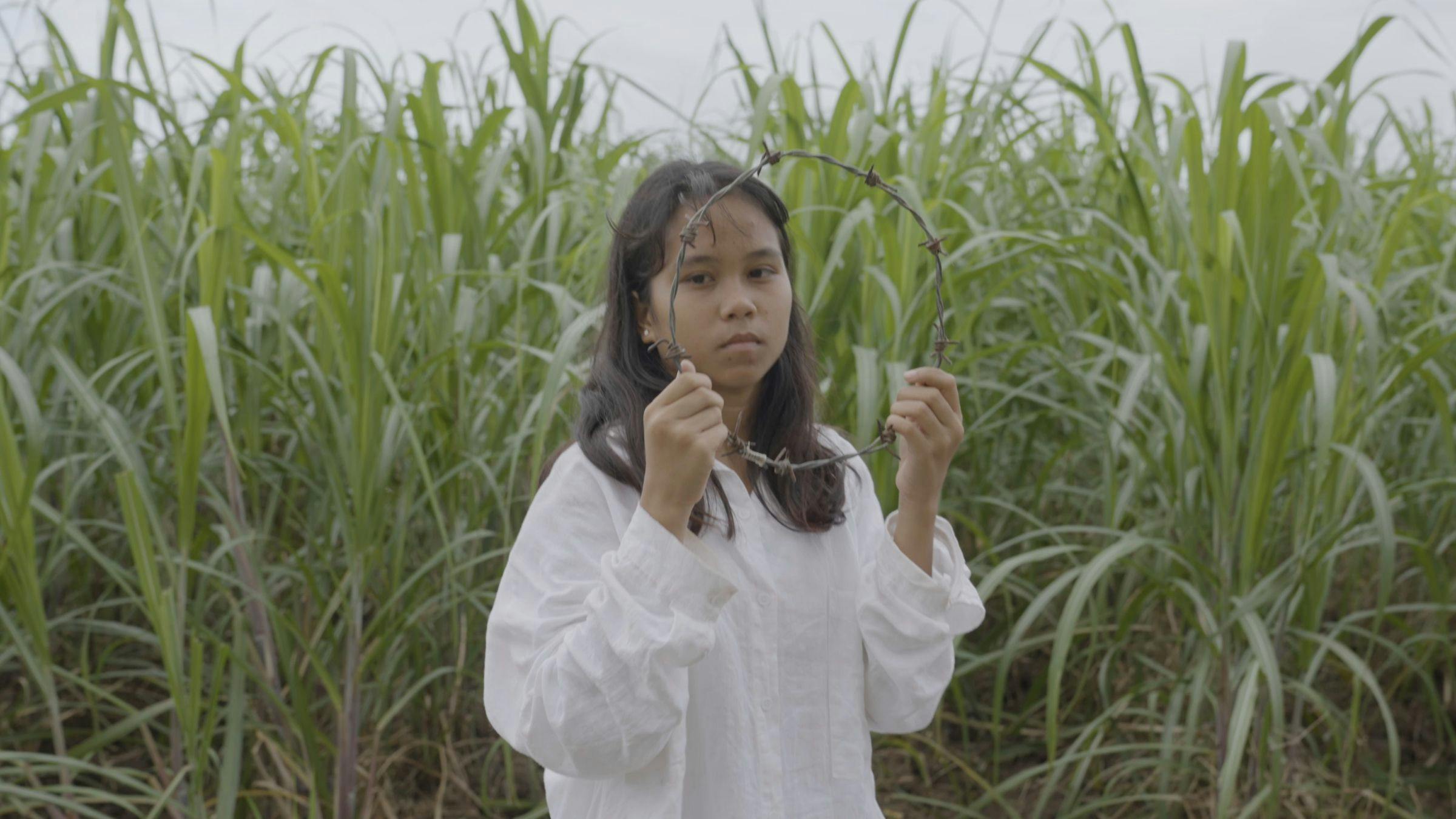 an image of a young person holding a circle of barbwire. She is wearing a white shirt and standing in a field of tall grass