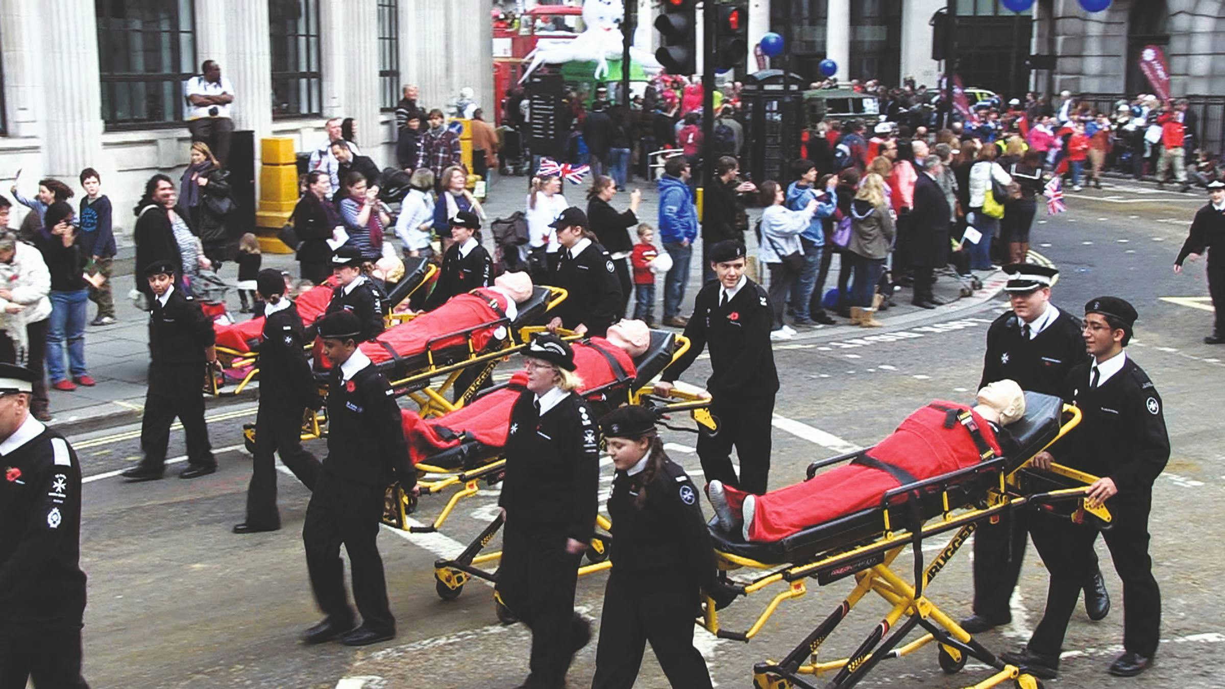 an image of mannequins being wheeled on stretchers by police people in a road. The pavement is lined with people