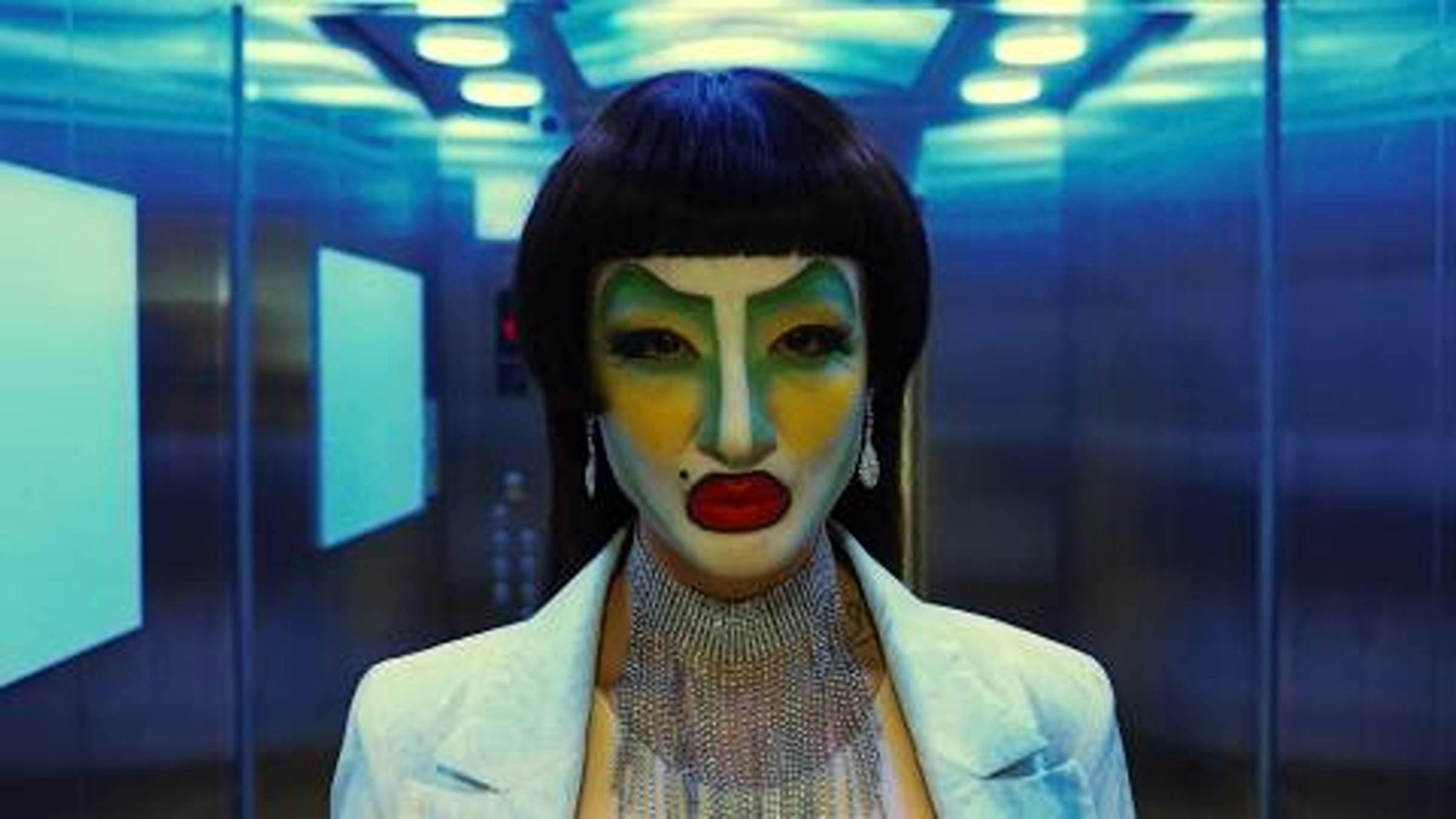 Image of a person looking directly at the camera wearing red lipstick and green and yellow face make up. The person is in a lift.