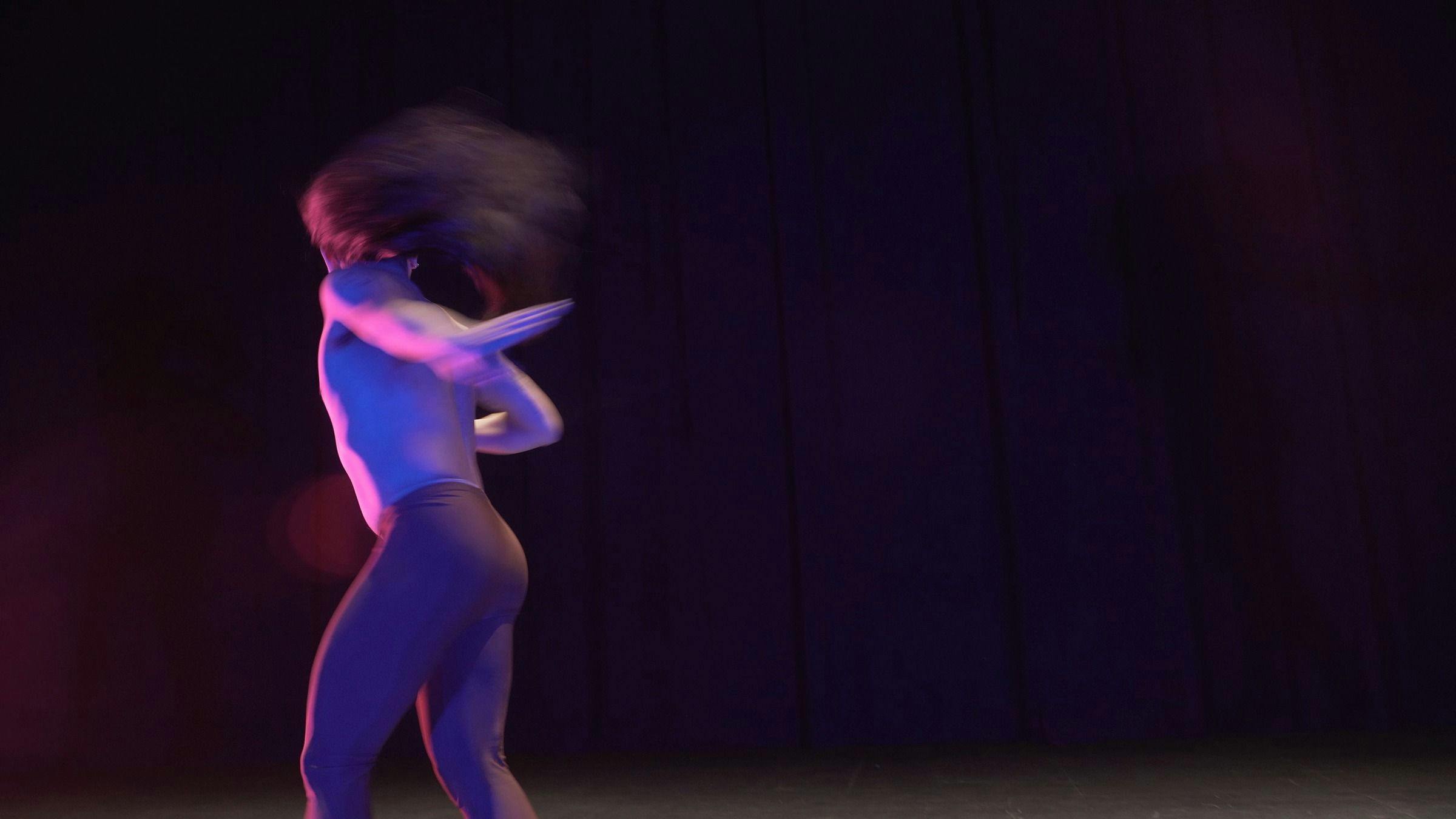a dancer who is wearing brown leggings spins in a darkened space. Purple light is cast on their body.