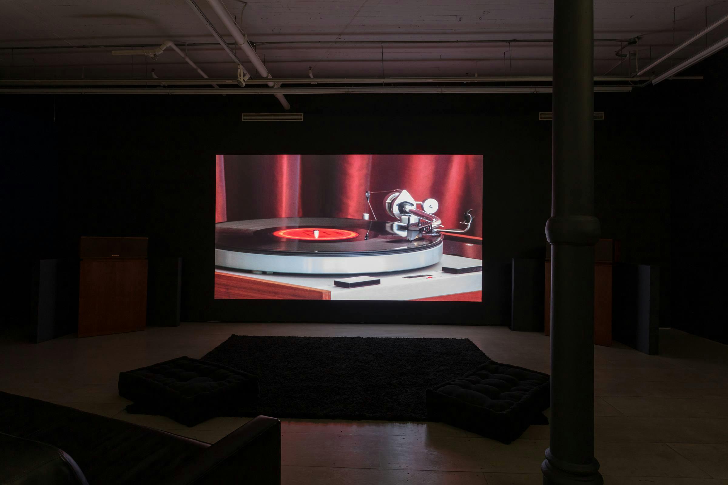 installation view of Last Night projected in a darkened gallery space. There are sofas in front of the projection and a concrete column is visible in the foreground
