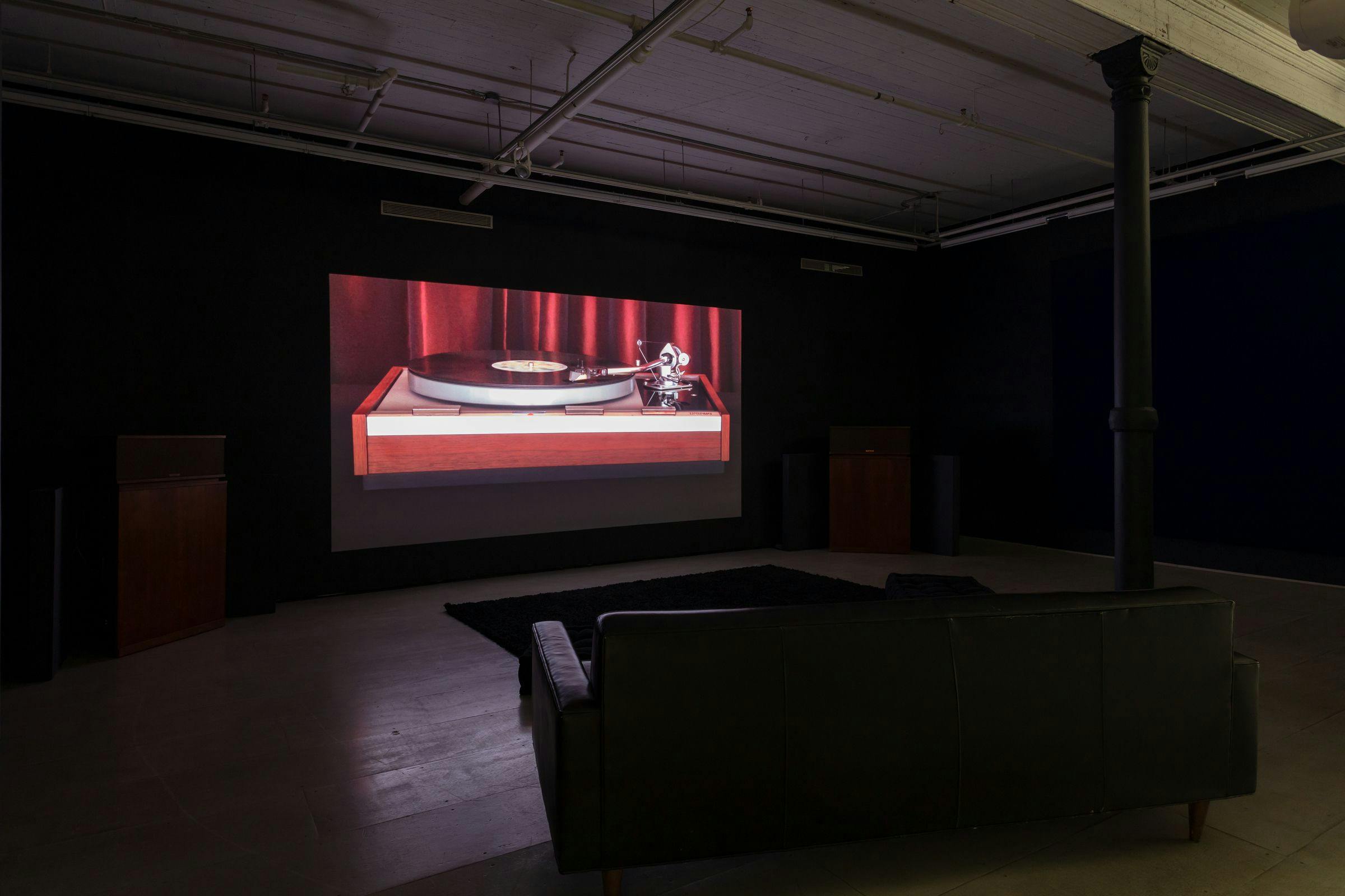 installation view of Last Night projected in a darkened gallery space. There are sofas in front of the projection and a concrete column is visible in the foreground.