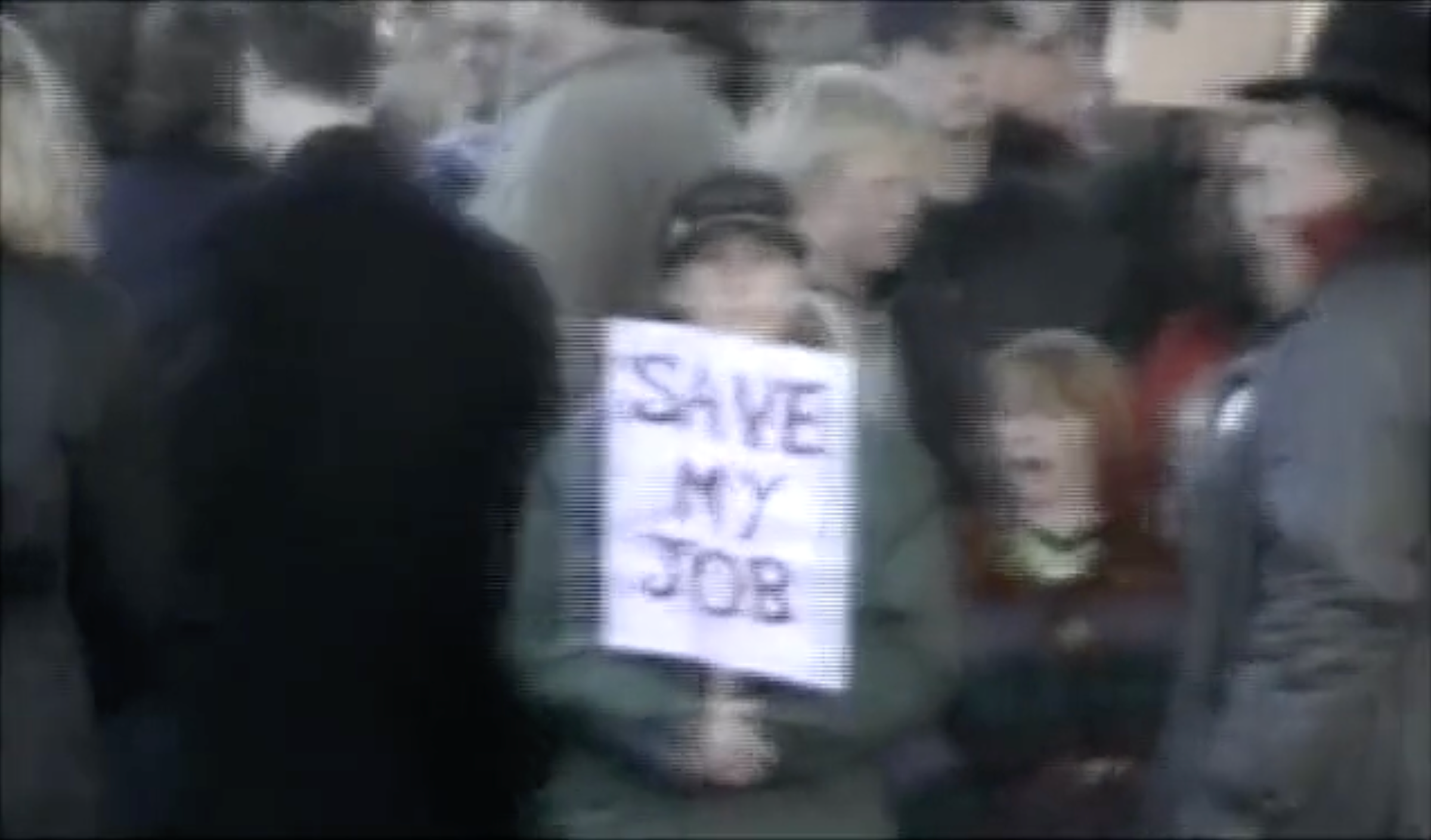 Shaky image of a crowd of people with one person looking forward at the camera holding a protest sign which reads 'SAVE MY JOB'.