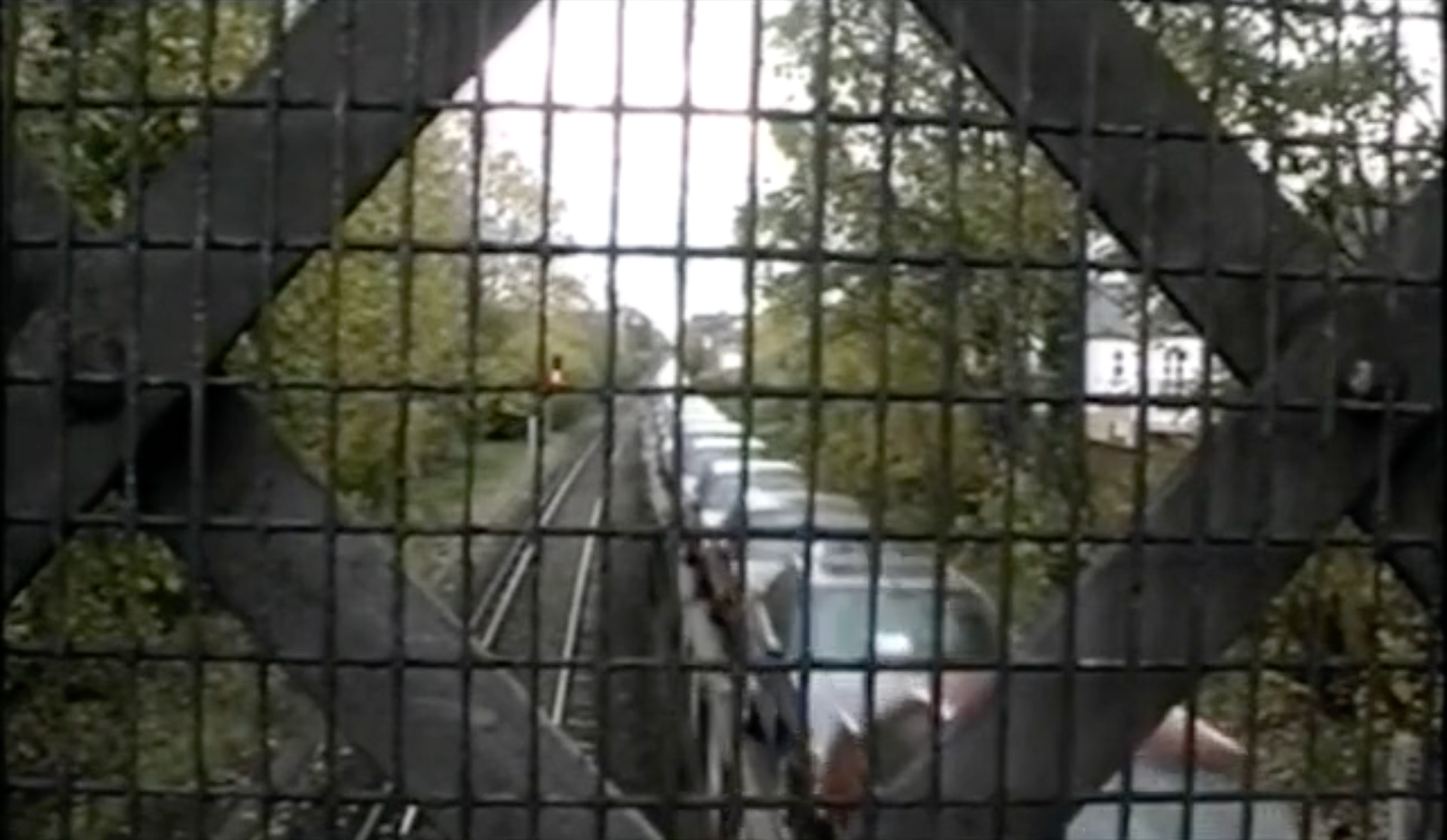 Image taken from a bridge looking down to train tracks below through a metal fence. A train goes past carrying cars.
