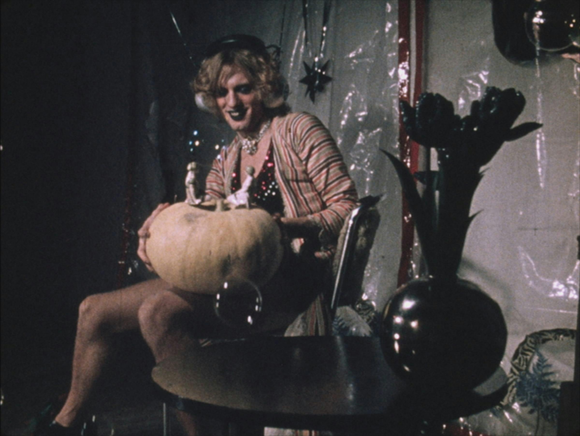 A femme person with blond curly hair sits at a circular table with a black vase on it. They hold a pumpkin with two figures on top.