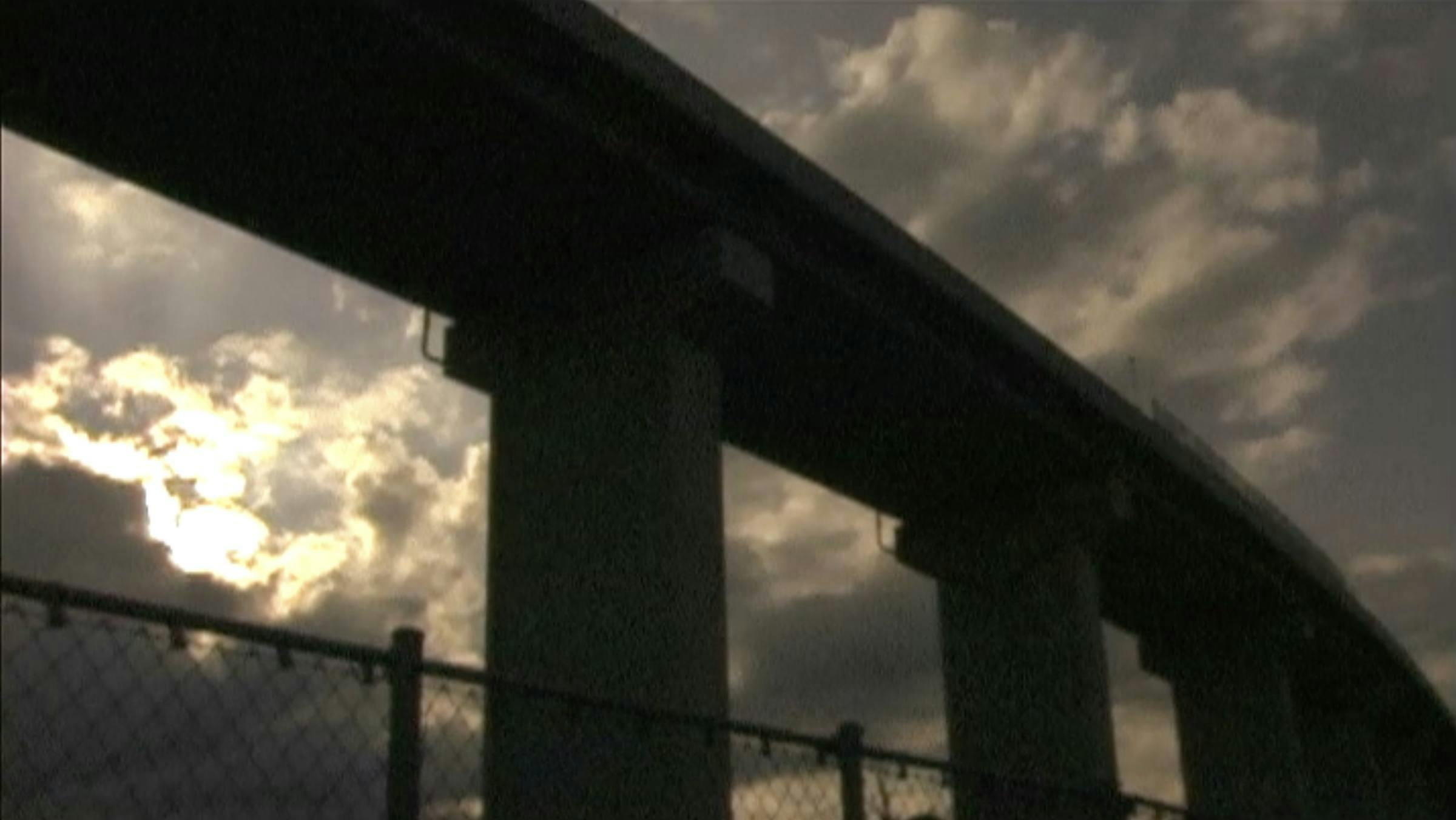 Image of a concrete underpass with chain link fence in the foreground, cast against a cloudy sky with the sun breaking through the clouds on the left.