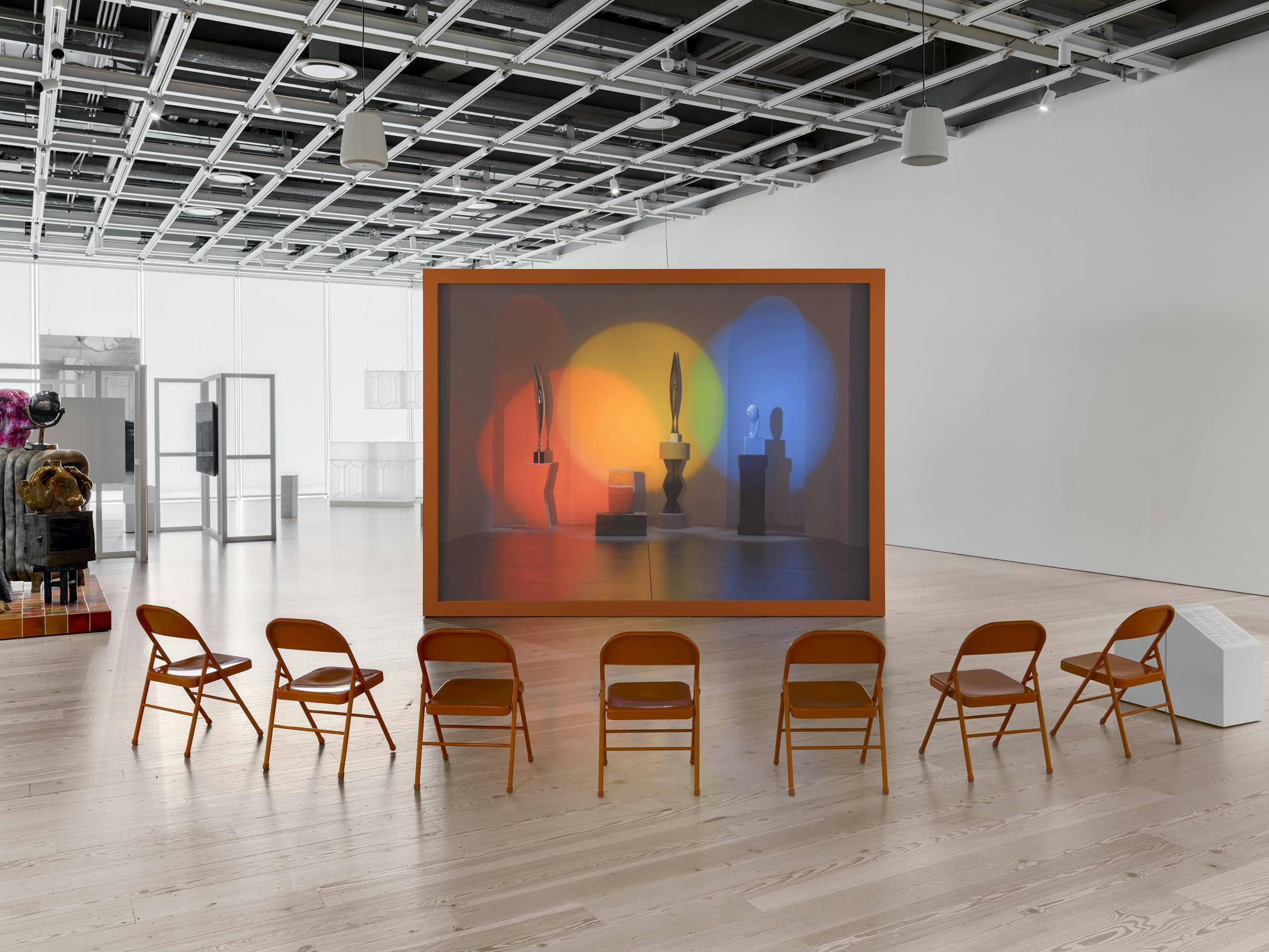 an art gallery with wooden floors and white walls. A video art work projected on to an orange freestanding box. 8 orange chairs are in front of the projection for the audience to sit on. 