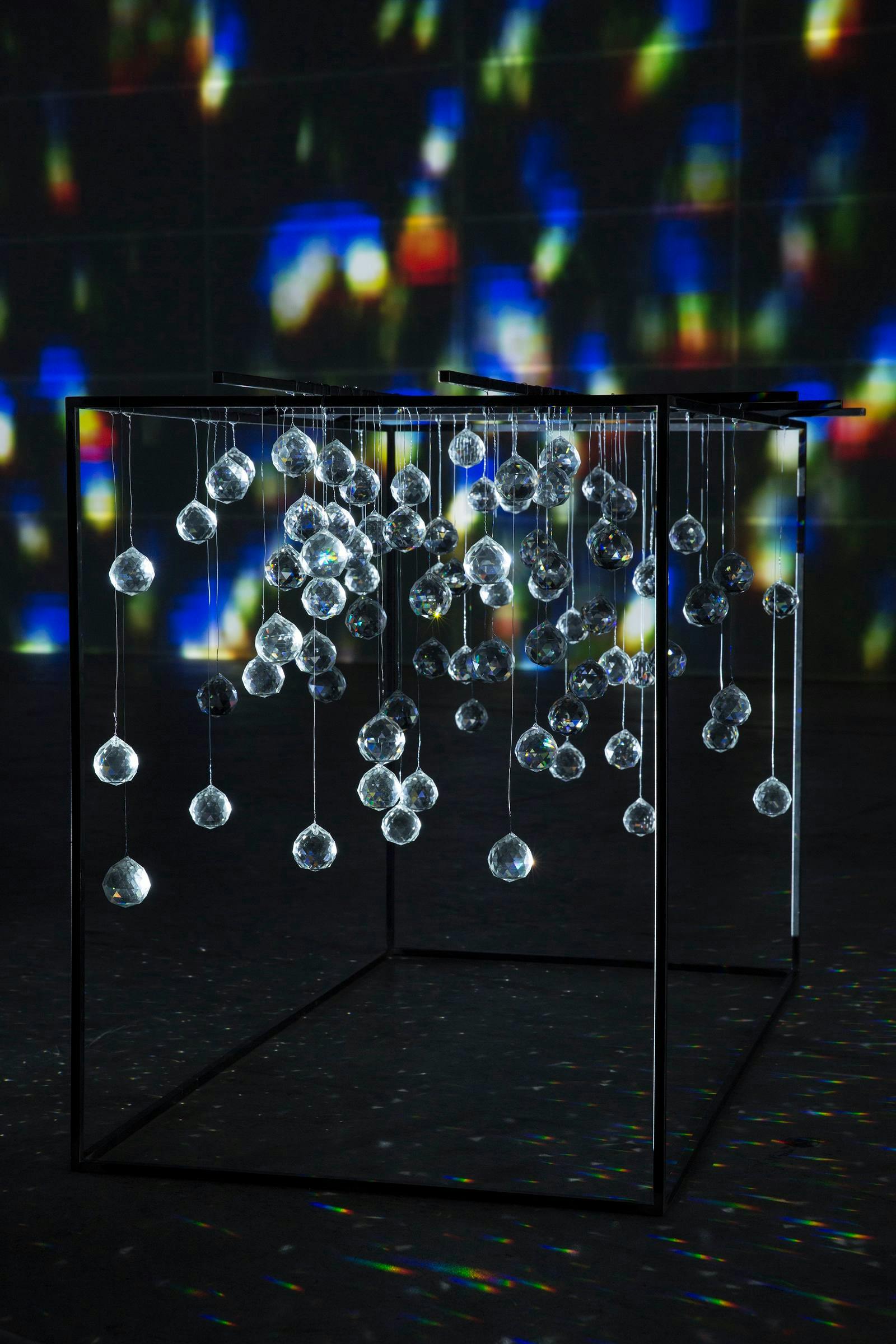 A metal open cube structure with glass spheres hanging from it in sits in a darkened space. The light refraction casts rainbow shadows on the walls