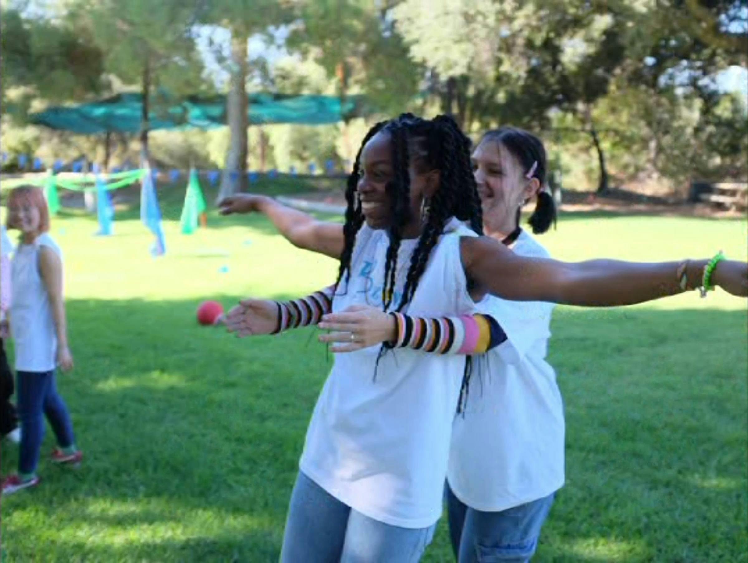 two young people wearing white tshirts are in a park. One person is leaning back on the other in anticipation they will catch her