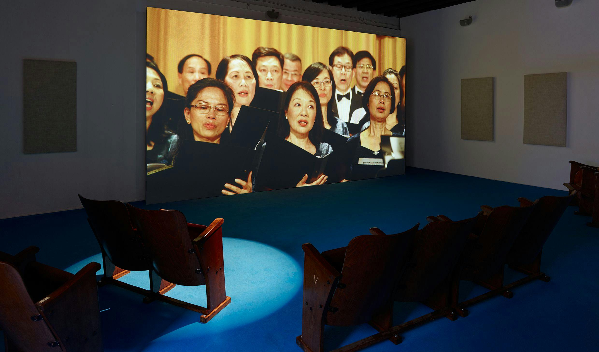 Installation view, Samson Young, We are the World, as performed by the Hong Kong Federation of Trade Unions Choir (Muted Situation #21) from Songs for Disaster Relief, Hong Kong Pavilion, 3 May - 26 November 2017, 57th Venice Biennale, Venice. We can see the work projected on the wall, beige sound panels and wooden church pews for the audience to sit on. The floor is covered in blue carpet.