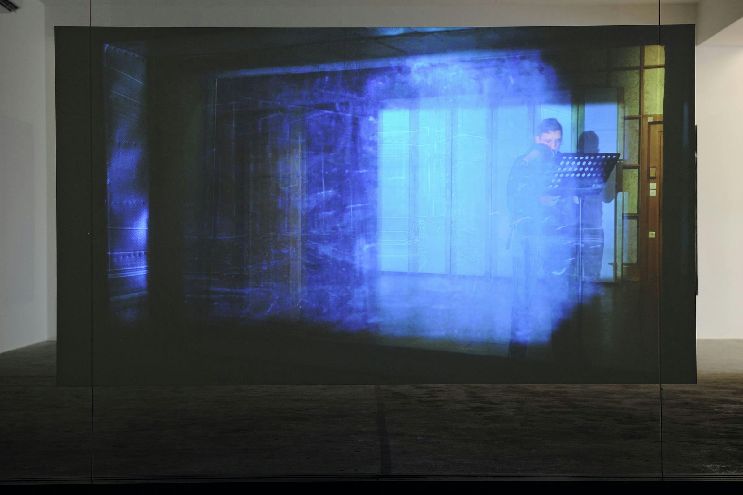 a projection of a video work in a gallery space which shows the artist who is wearing glasses standing in front of a lectern. 