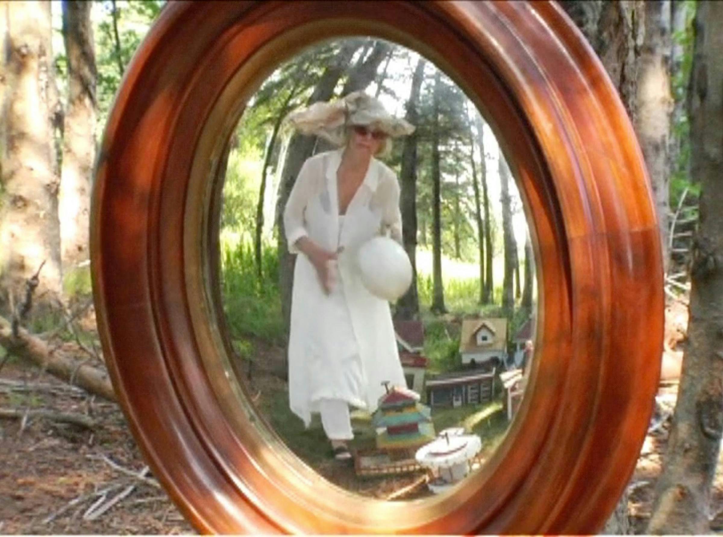 An image of the artist dressed in white and standing in a garden. She is wearing sunglasses. She is peering through a wooden window at us