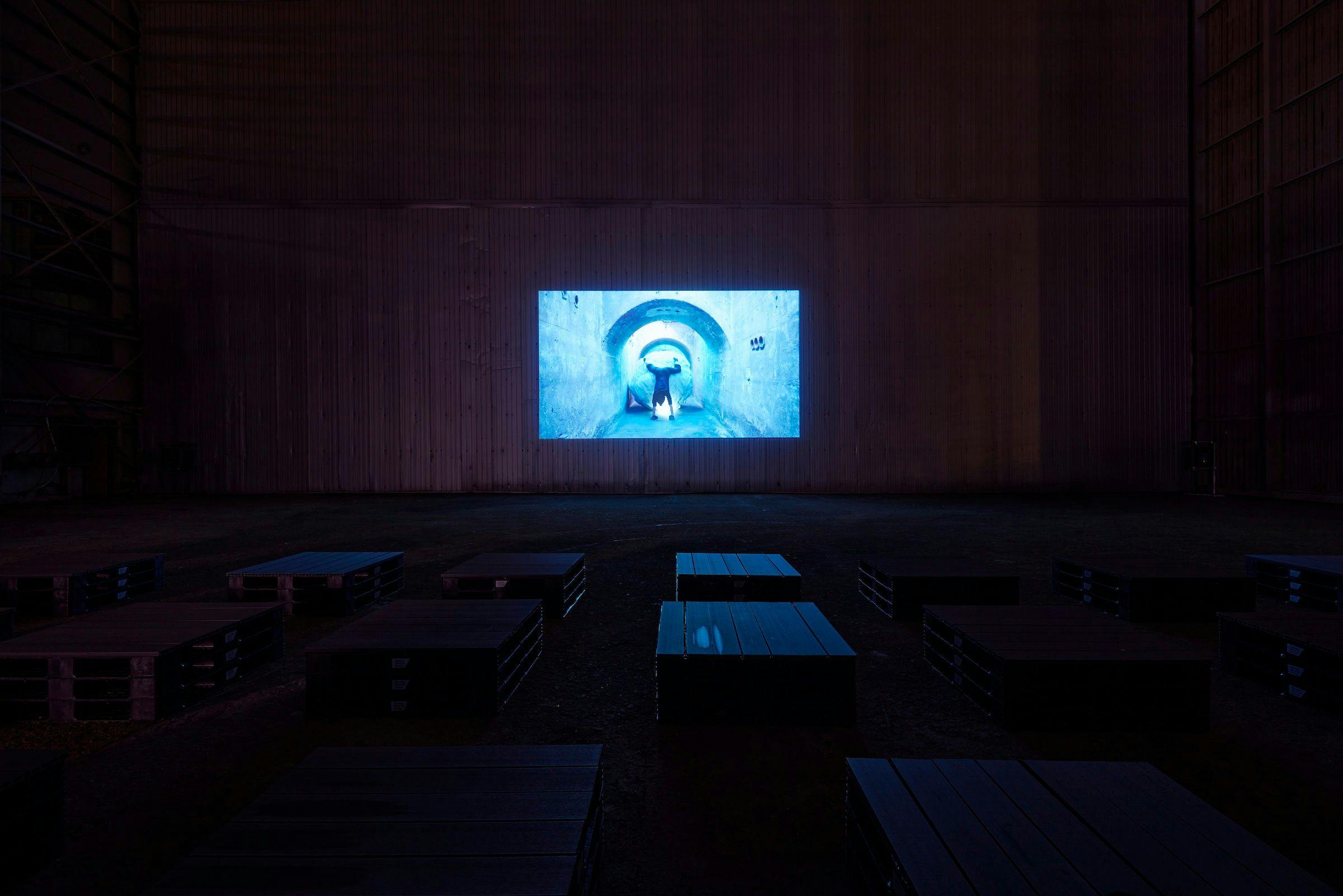 an installation of a video projection in an art gallery. The walls and floor are brown and there are a number of plan chest draws in front of the projector