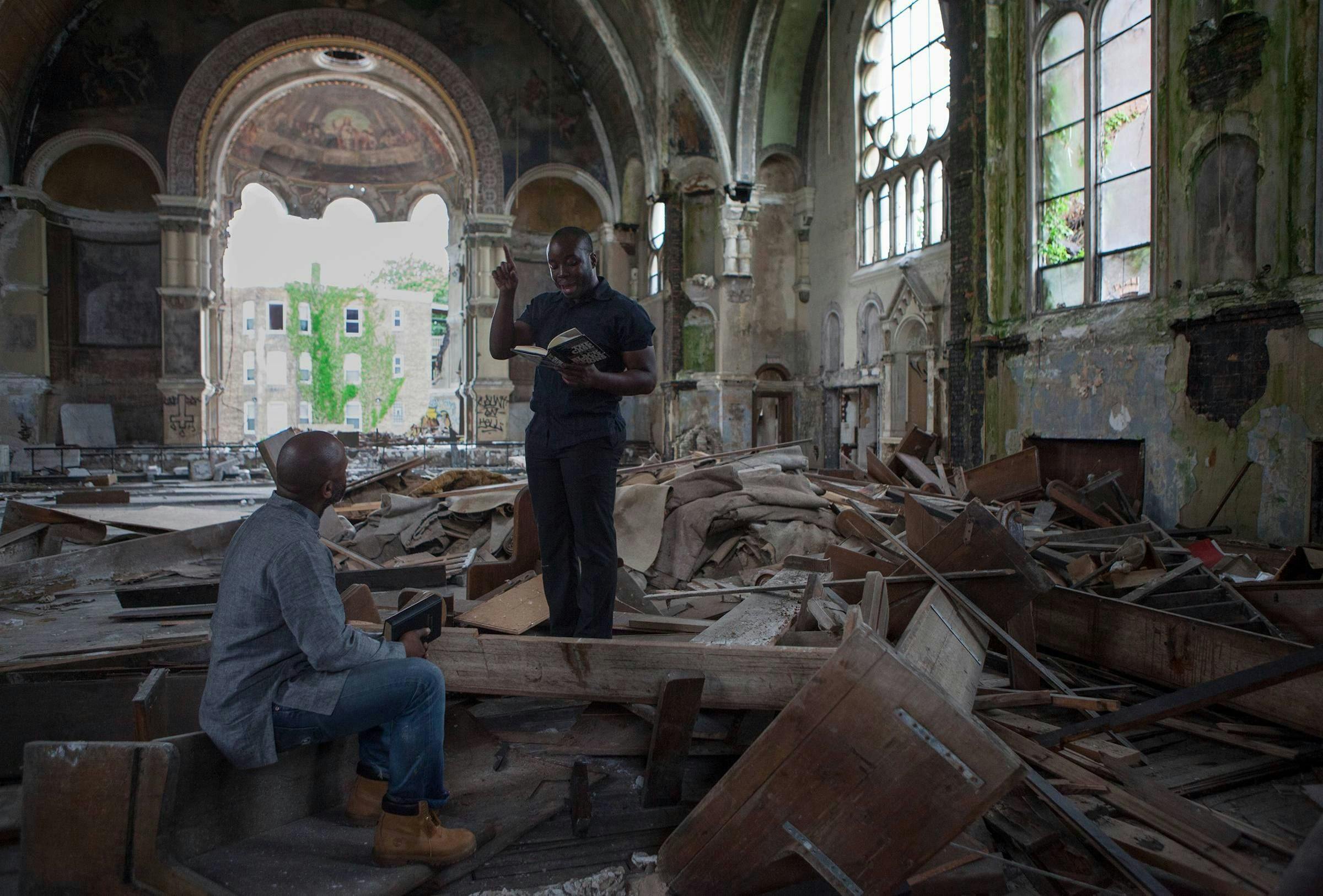 Images of the original performance and demolition of the church. One black monks stands among the ruins, the other is seated. The standing person is reading a loud from a book they hold in their hands