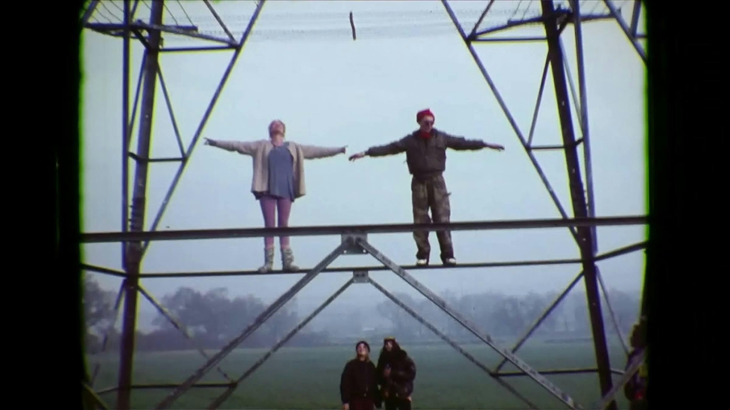 an image of 2 peole standing on a metal rung of an electrical pylon in a field. They have both of their arms out stretched. Two people on the ground are looking up at them