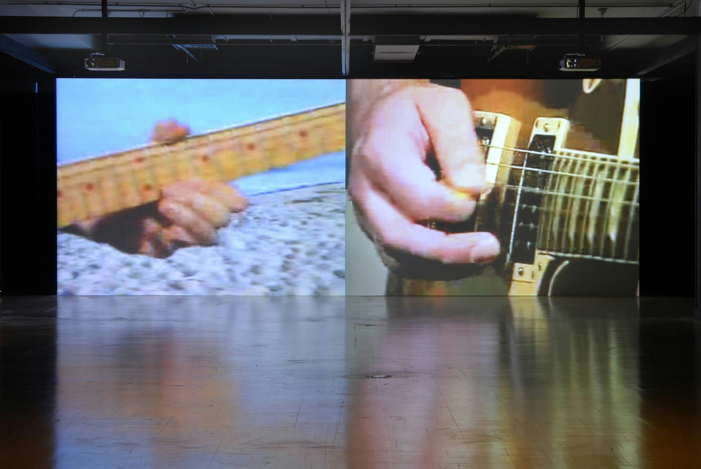 Image of a gallery setting with two large projections of people's hands playing a guitar next to one another