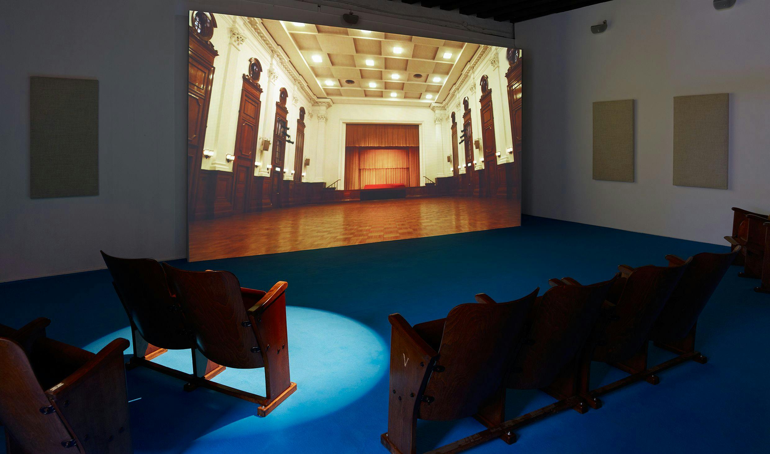 Installation view, Samson Young, We are the World, as performed by the Hong Kong Federation of Trade Unions Choir (Muted Situation #21) from Songs for Disaster Relief, Hong Kong Pavilion, 3 May - 26 November 2017, 57th Venice Biennale, Venice. We can see the work projected on the wall, beige sound panels and wooden church pews for the audience to sit on. The floor is covered in blue carpet.