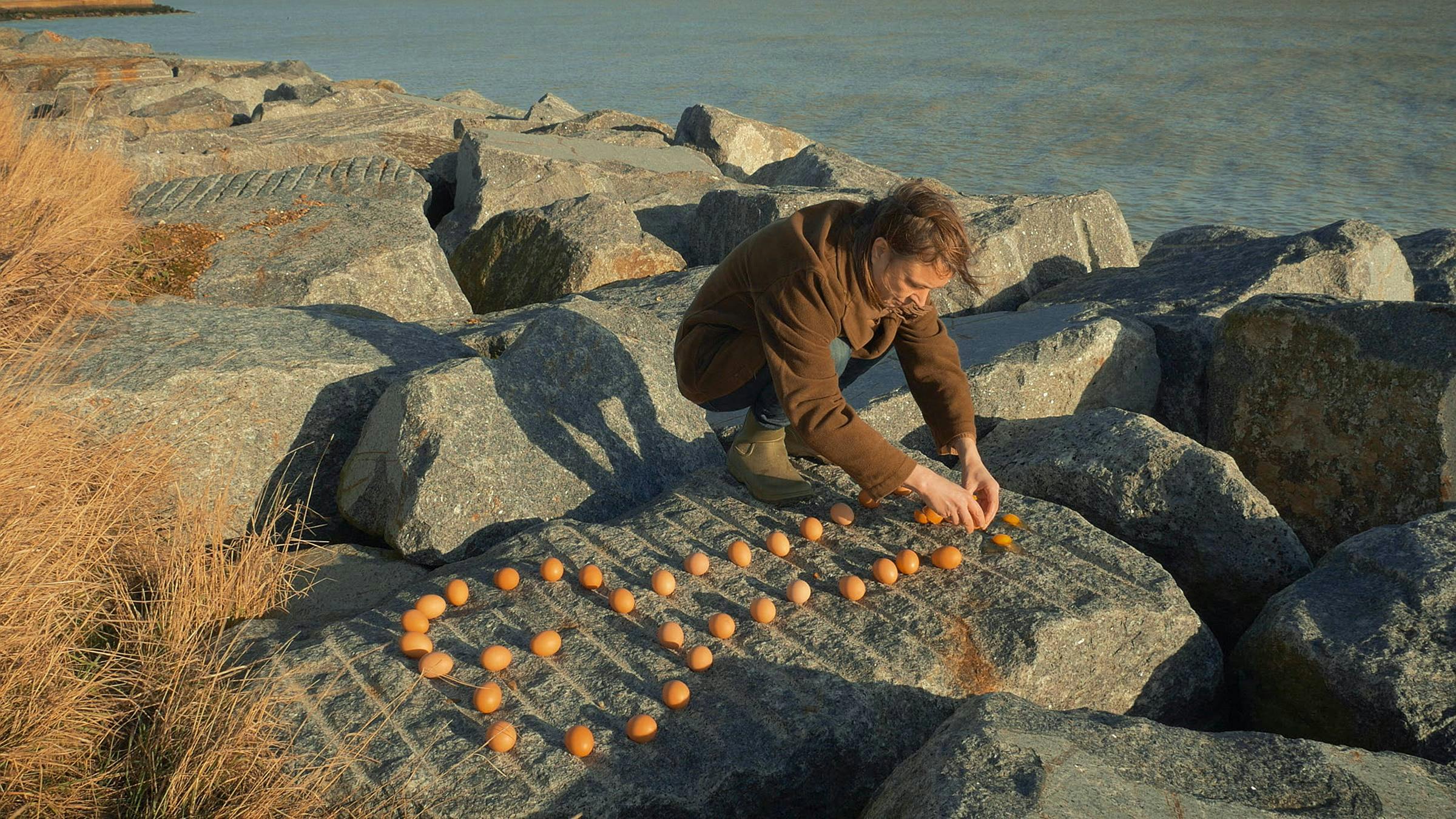 A person wearing brown clothes arranges lots of eggs on a rock in front of the ocean.