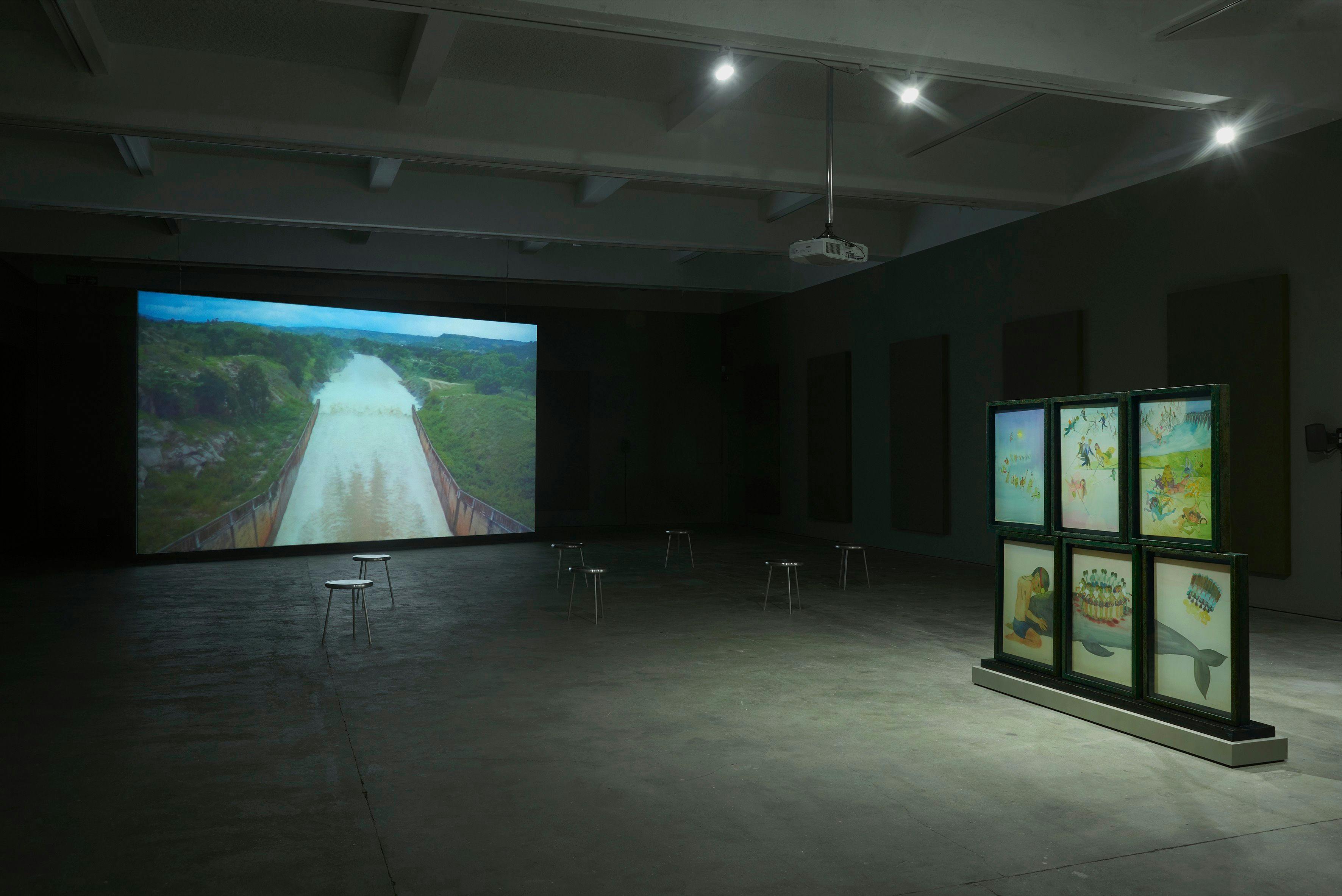 Image of a gallery space with a large screen on the left and six smaller screens pushed together on the right. There are lots of stools dotted around the space.