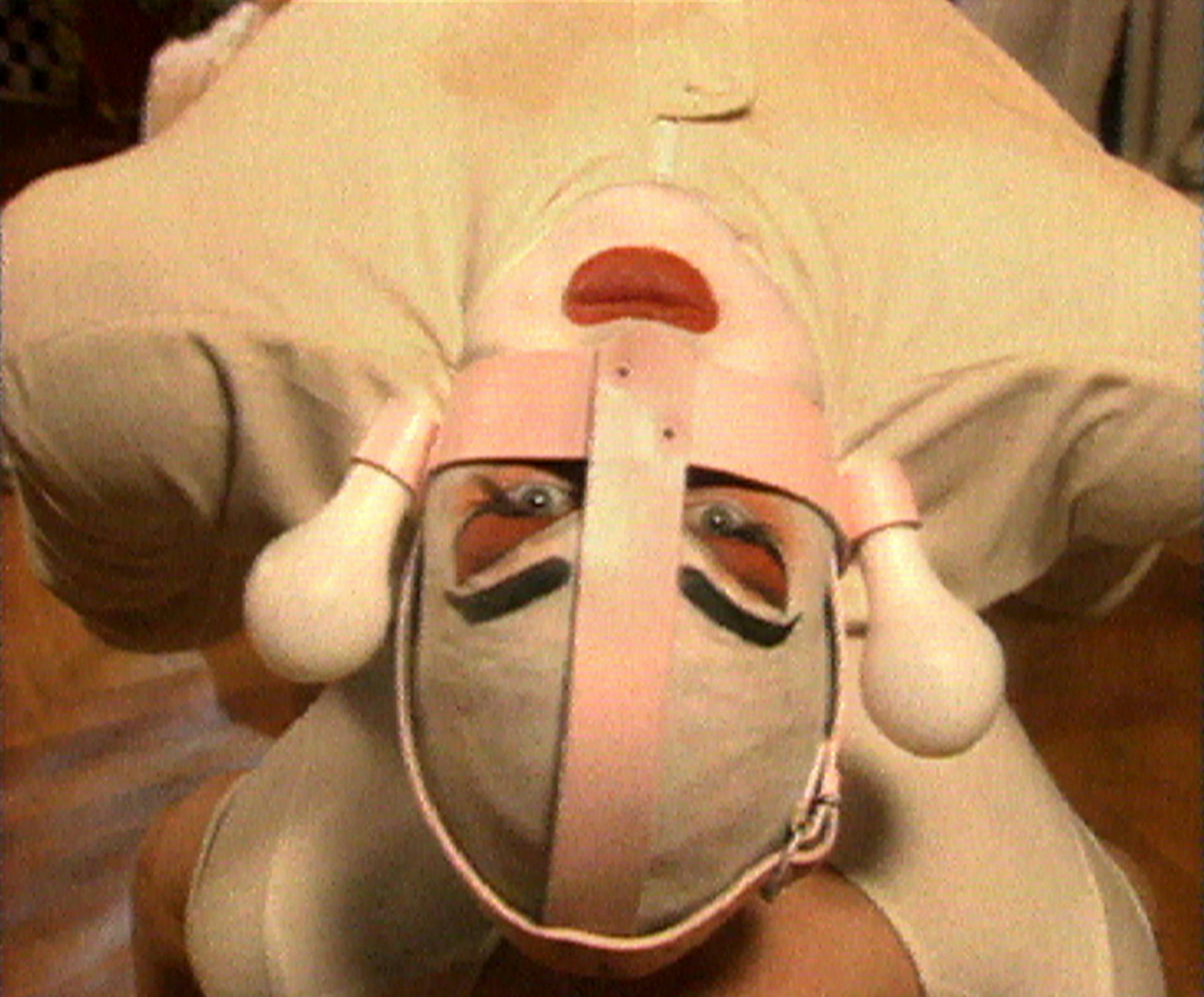 An image of a person leaning backwards. They are wearing a white body suit, their face is painted white and they gave pink leather straps wraped over their face in a cross shape. There are opaque white light bulbs either side of their face.