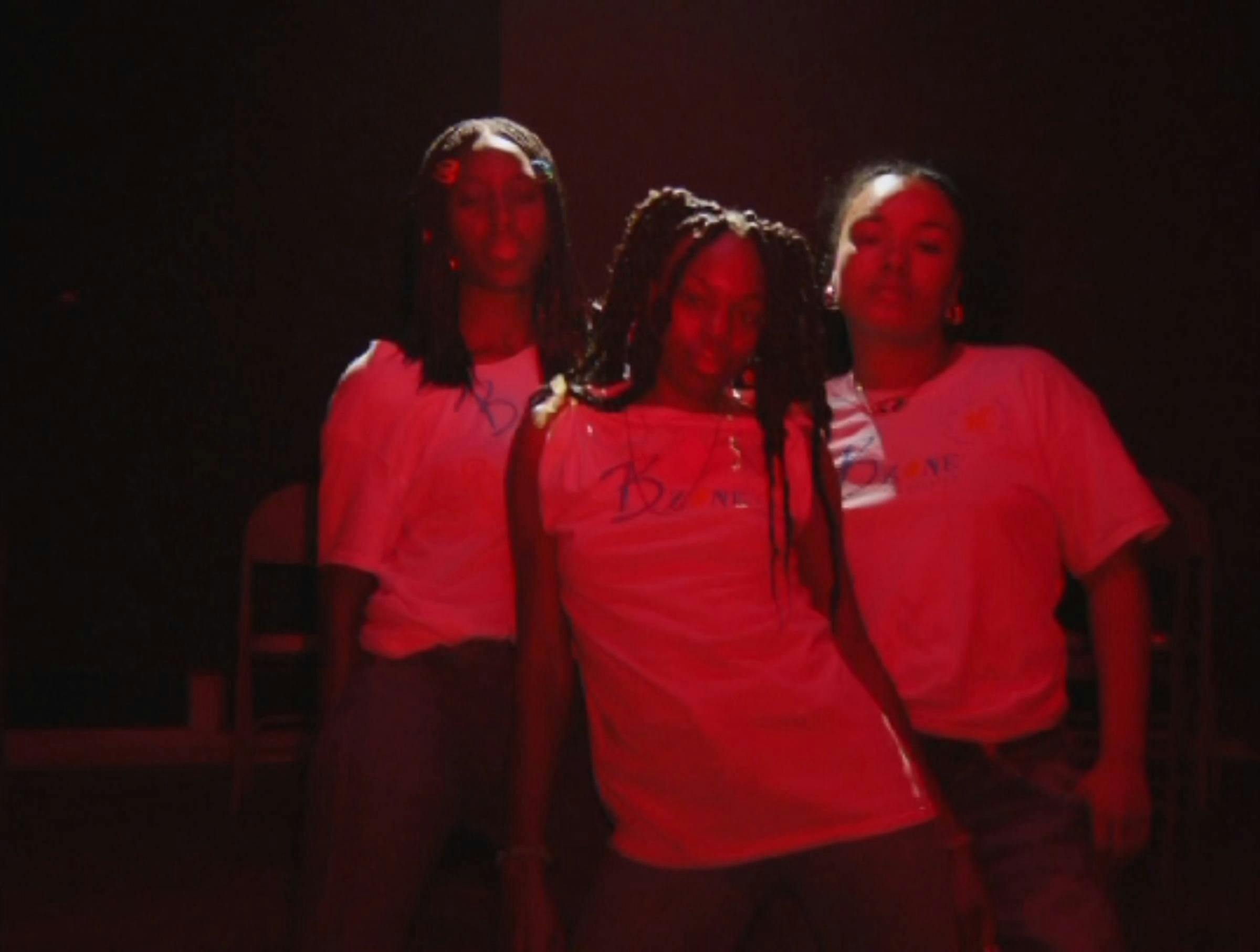 Three young black women stand in a row. They are posing. They are standing in red light which tints the whole image.