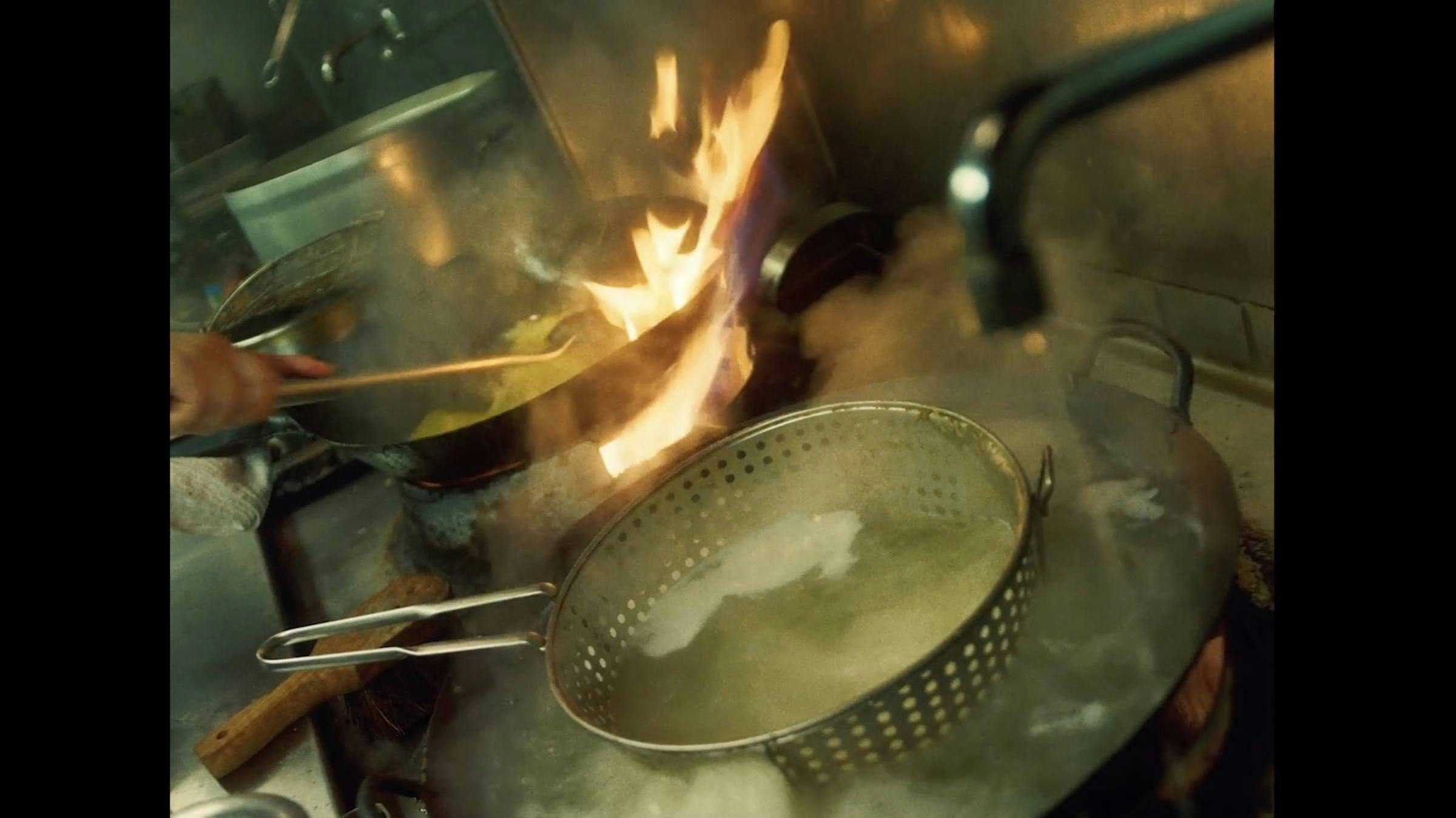 a busy resturant kitchen scene. We can see a pan on fire and a colander being submerged in a sink 