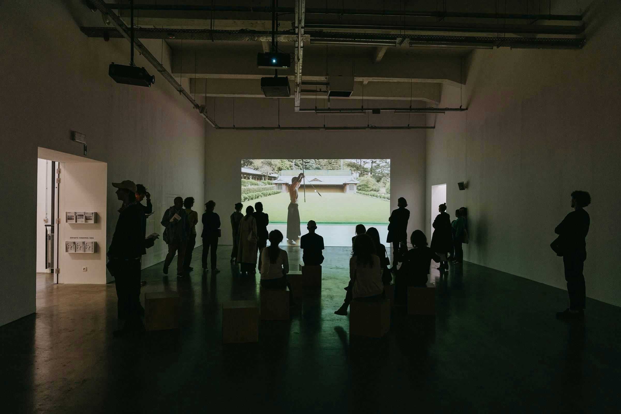 Gallery space with lots of people sat or standing, watching a video projected onto the far wall.