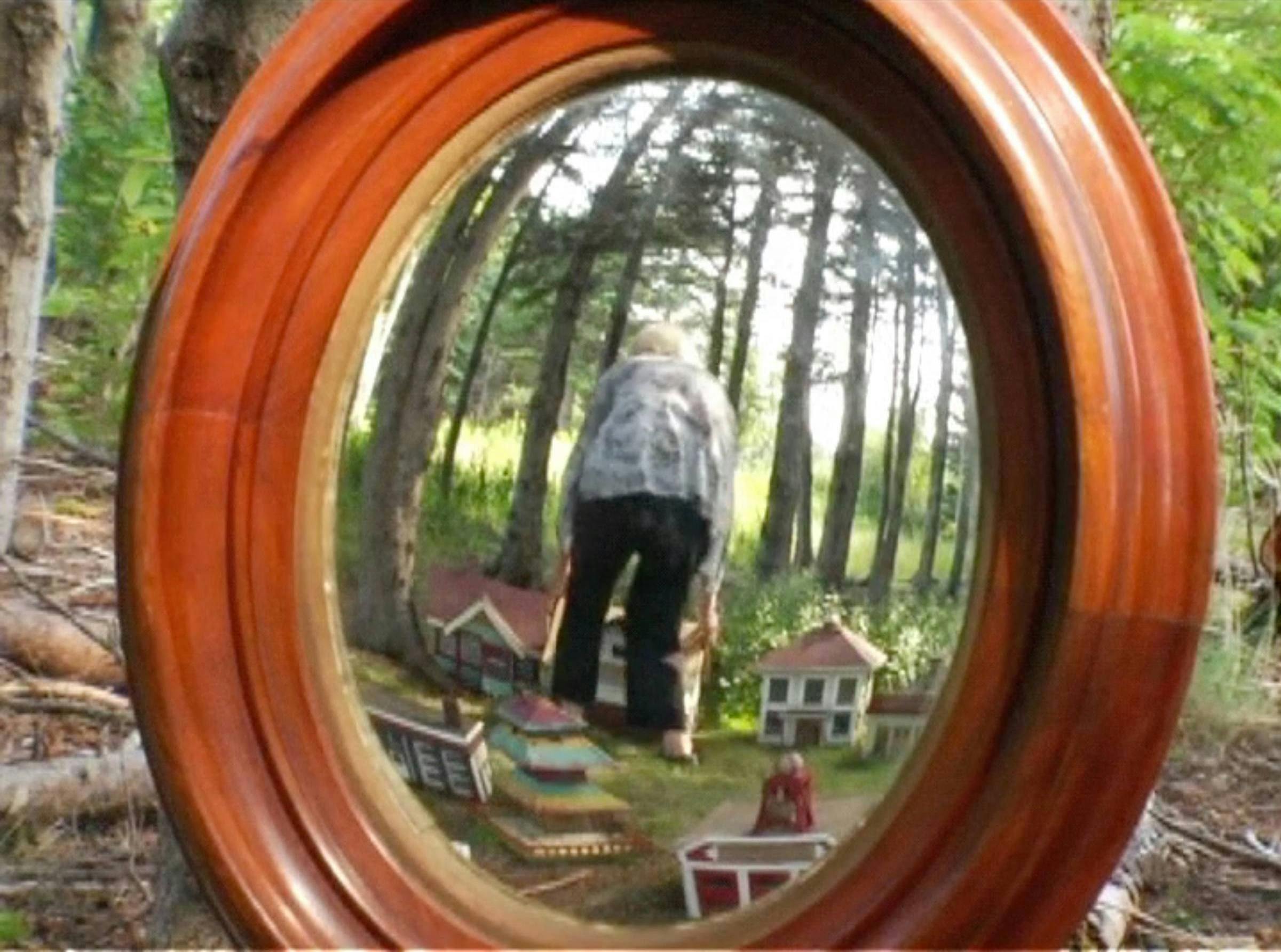 An image of the artist in a garden. She is peering through a wooden window at us. There are small wooden houses (doll size) in the garden