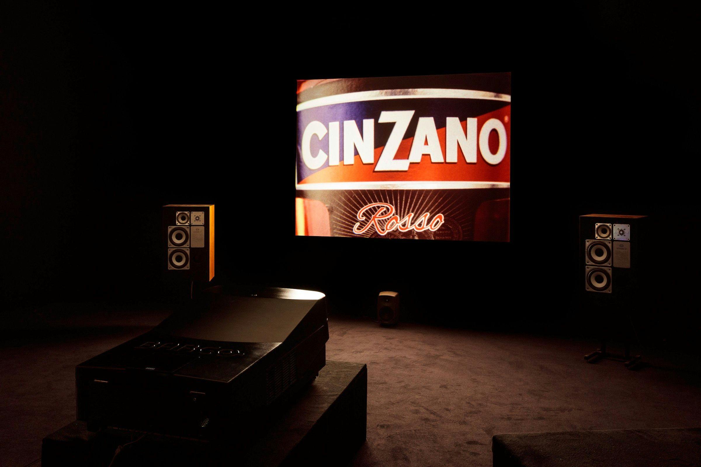 an installation view of Dream English Kid, 1964-1999 AD. The image of the work projected is a label from a Cinzano bottle. There are speakers either side of the projection and the projector is visible