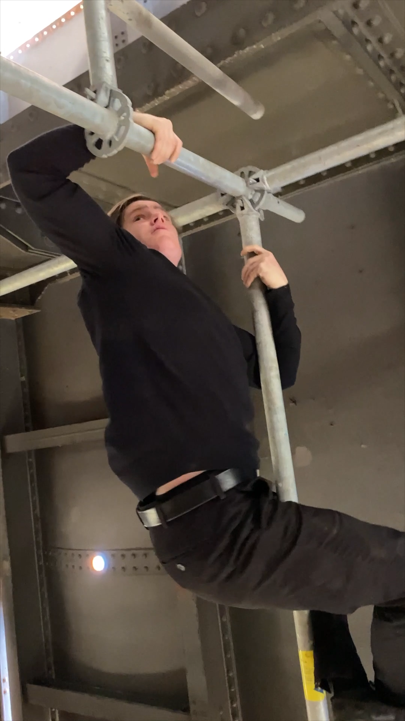 Person wearing a black outfit climbing on some scaffolding situated indoors.