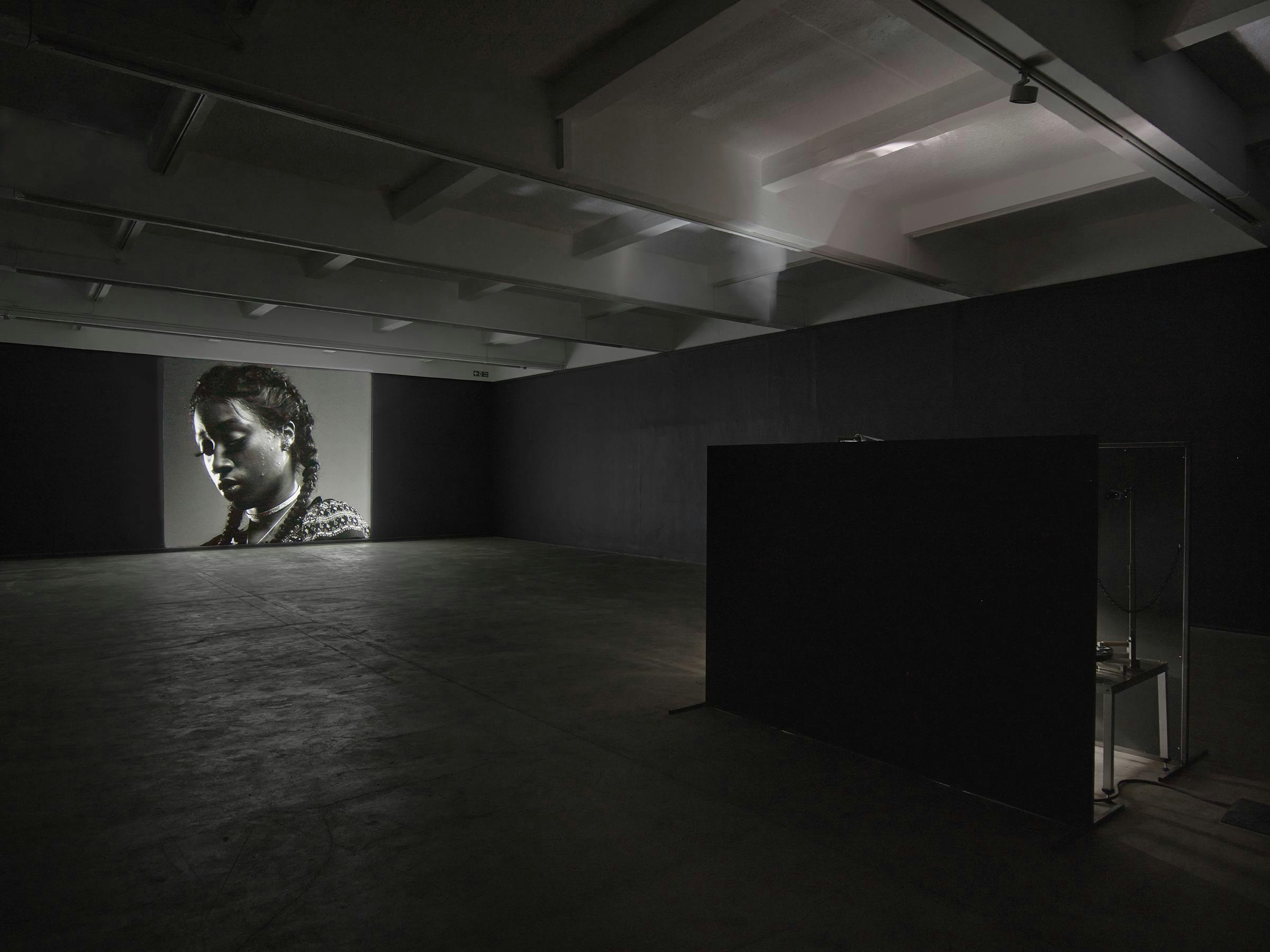 A video art work projected in a darkened space. The projected image is black and white and depicts a black woman wearing glasses and a white shirt at a 3 quarter length profile. We can see the projection box in the foreground.