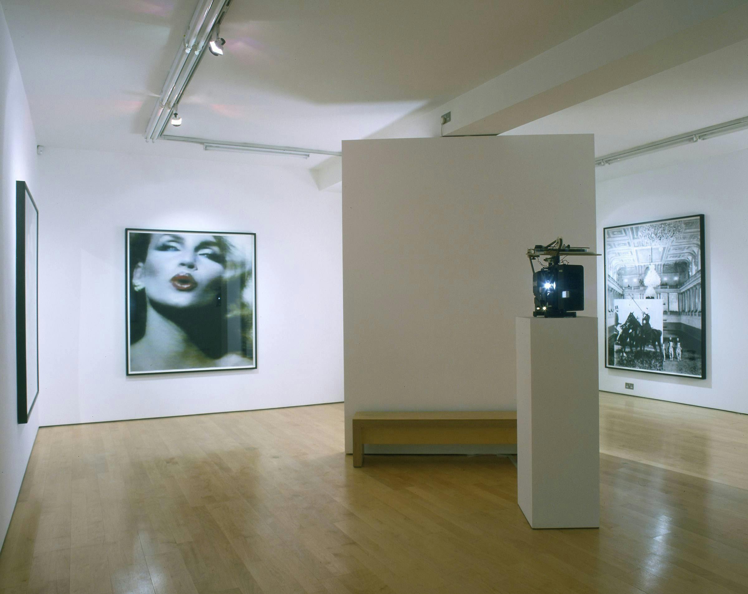 Image of a gallery where three large works hang on the wall and there is a bench and object on a plinth