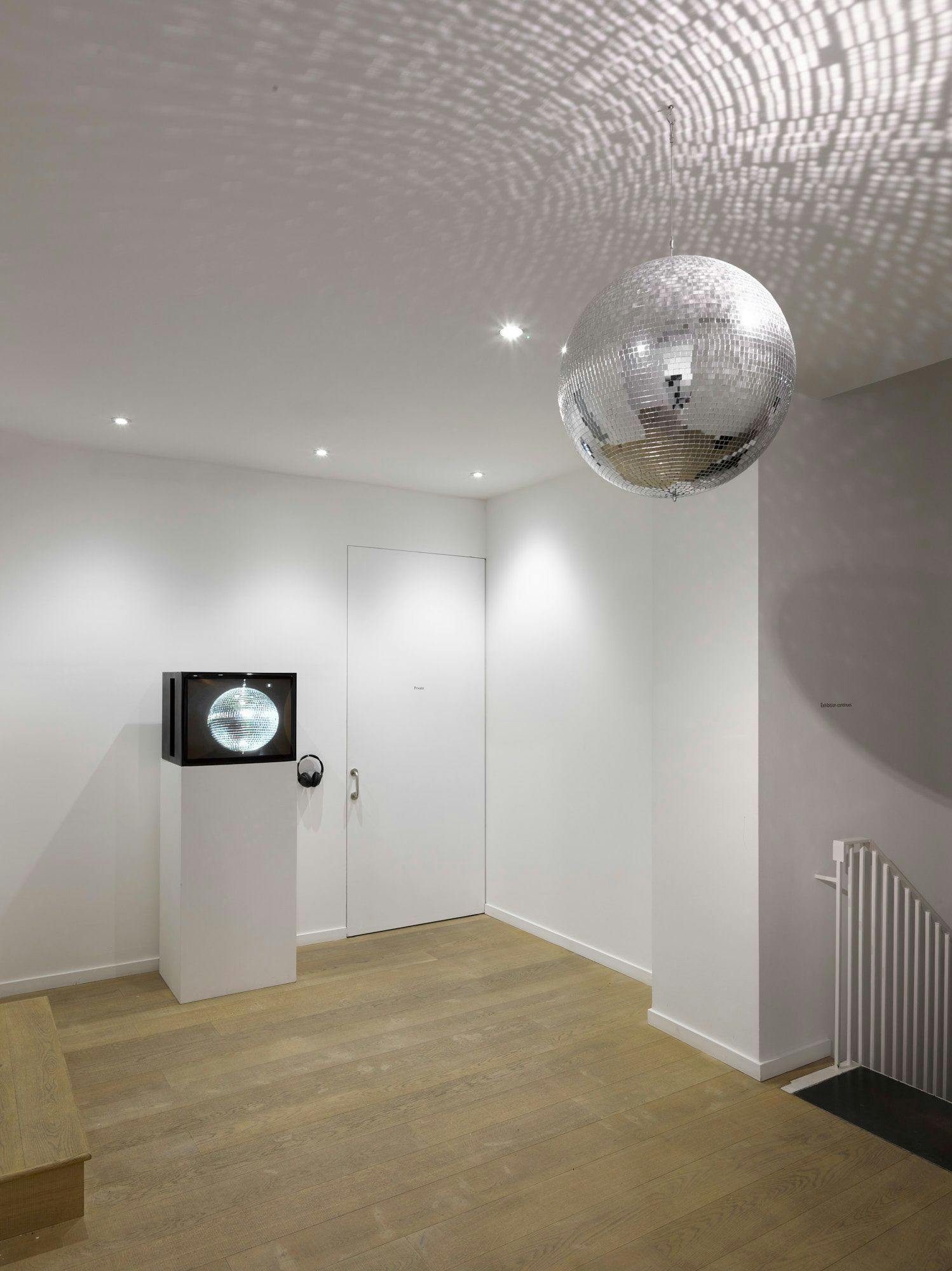Image of a white painted gallery space with wooden floors with a disco ball hanging from the ceiling and a TV on top of a plinth.