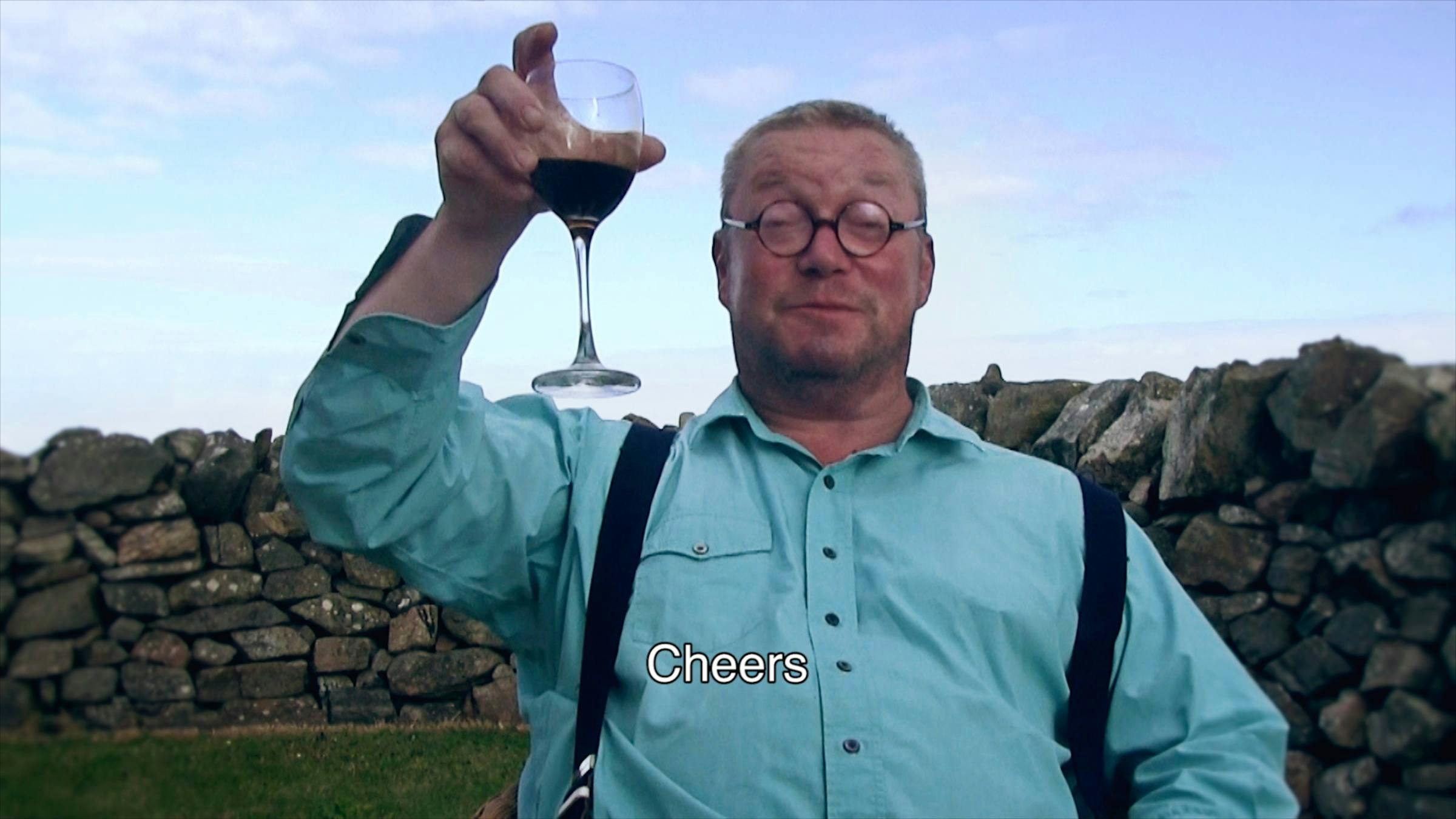 A man wearing a blue shirt with braces on top, small circular glasses, holding up a glass of red wine. In the background there is a stone wall and grass. The word 'Cheers' is superimposed on top.