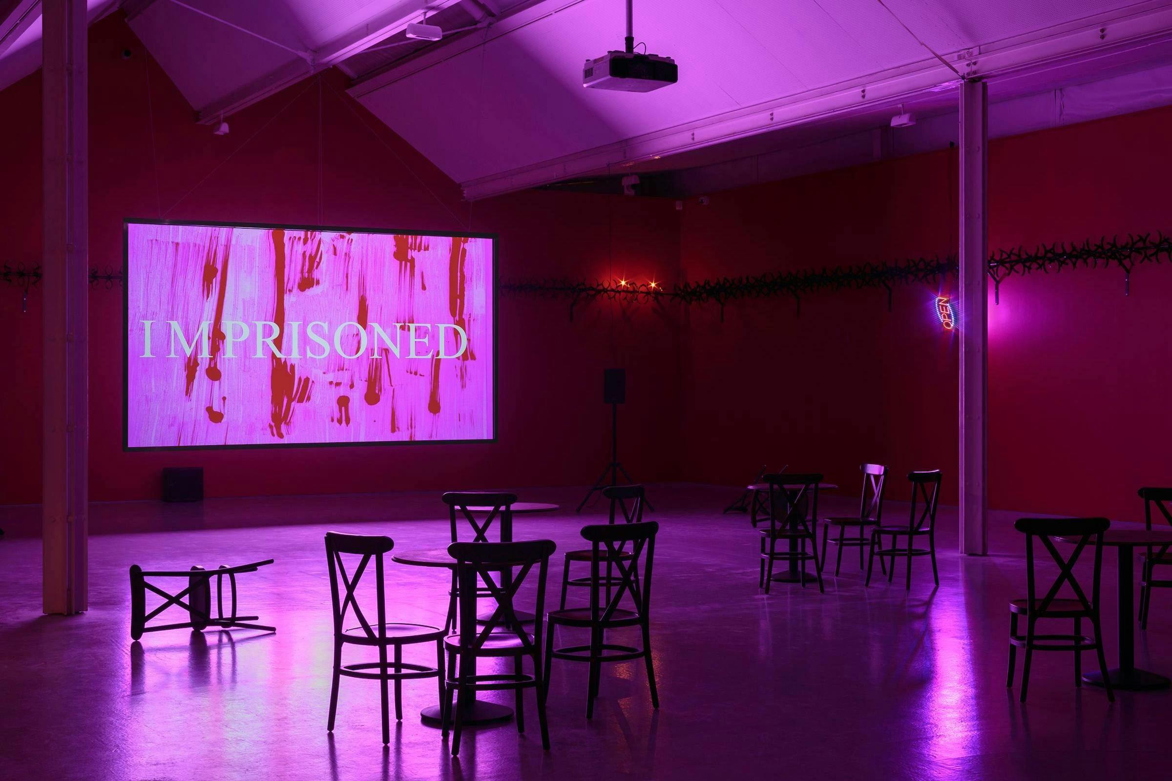 A video art work installed in a gallery space. There are a number of chairs and tables in front of the projection. The room is tinted with pink light.