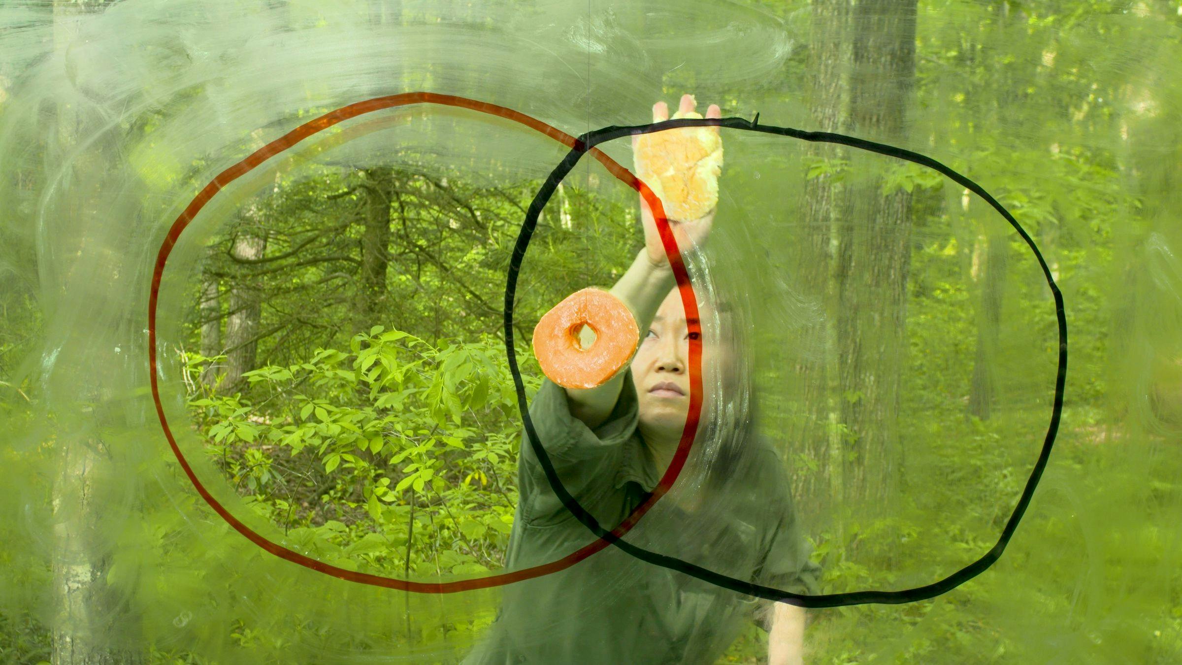 an image of the artist in a forest drawing a red circle and a black circle (on the camera lens) that overlap in the middle. In the section that overlaps we can see a doughnut