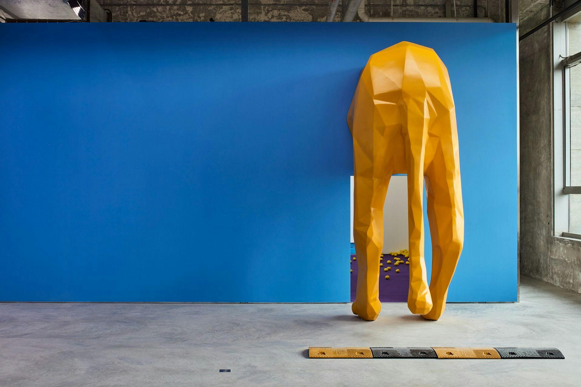An installation view of Installation view, Samson Young, The highway is like a lion's mouth, 6 November - 23 December 2018, Edouard Malingue Gallery, Shanghai. We see a blue wall with a door way. We can see the back side of a giant yellow lion. People need to walk under the lion to enter the room behind