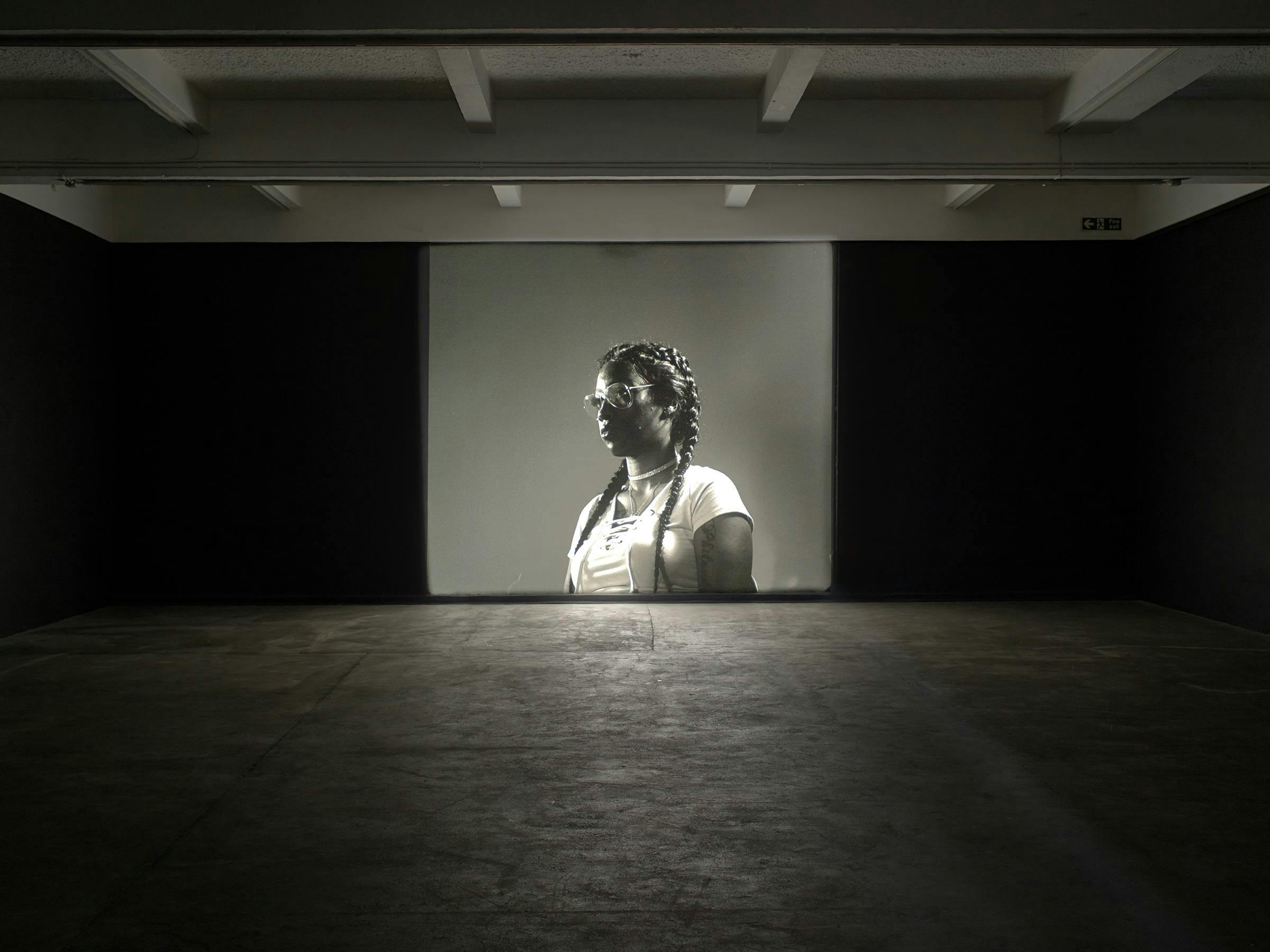 A video art work projected in a darkened space. The projected image is black and white and depicts a black woman wearing glasses and a white shirt at a 3 quarter length profile.