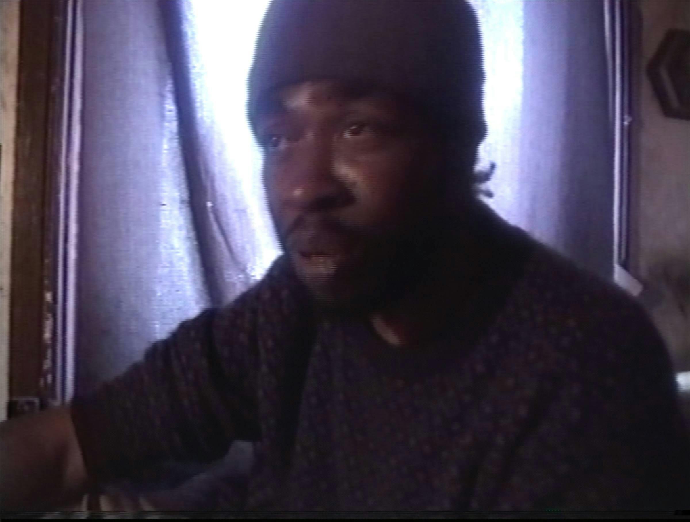 Image of a man wearing a dark jumper and beanie sat in a room with a window behind him covered by a piece of material