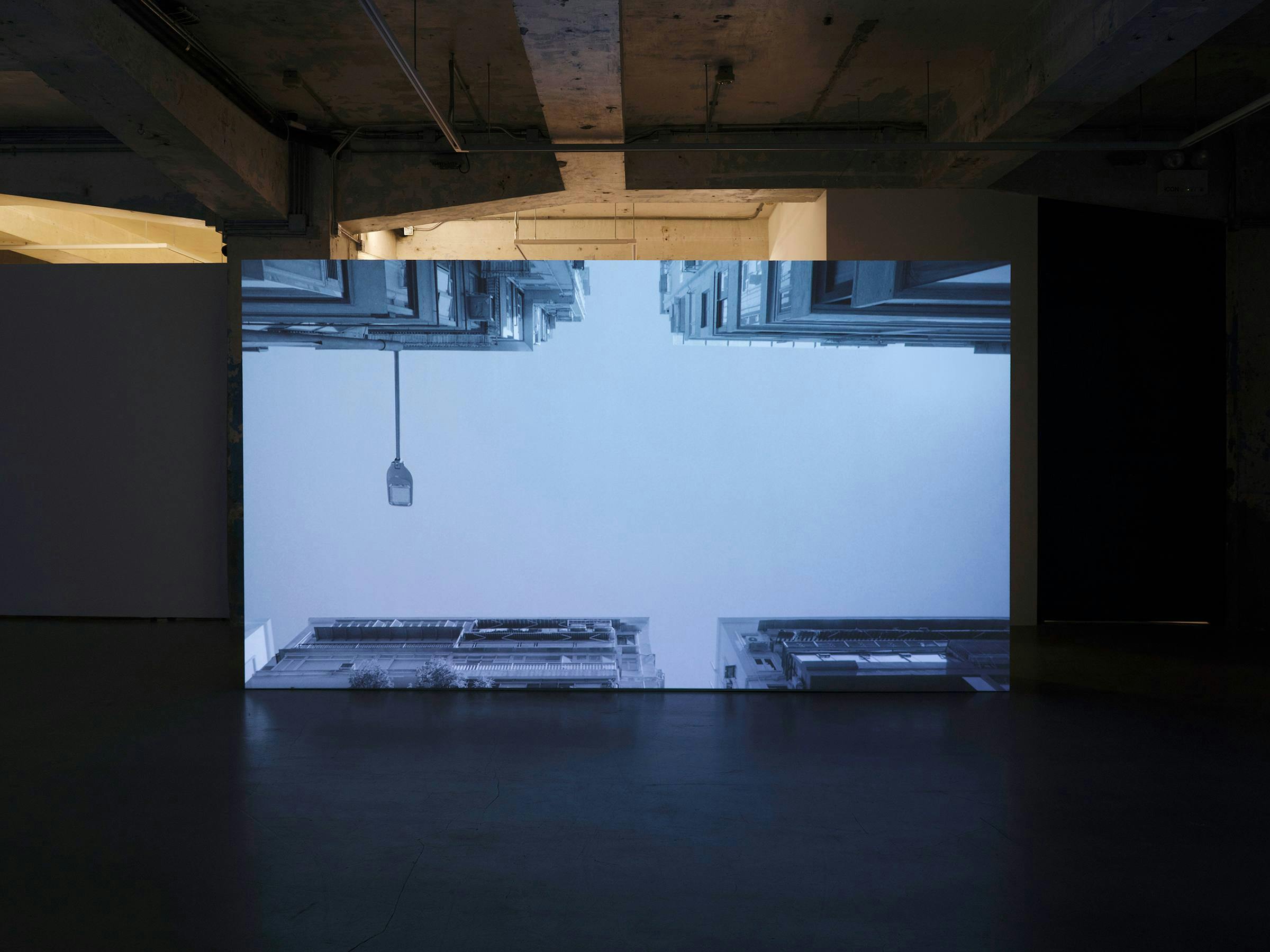 Large projection of a black and white image of buildings and sky in a concrete gallery space.