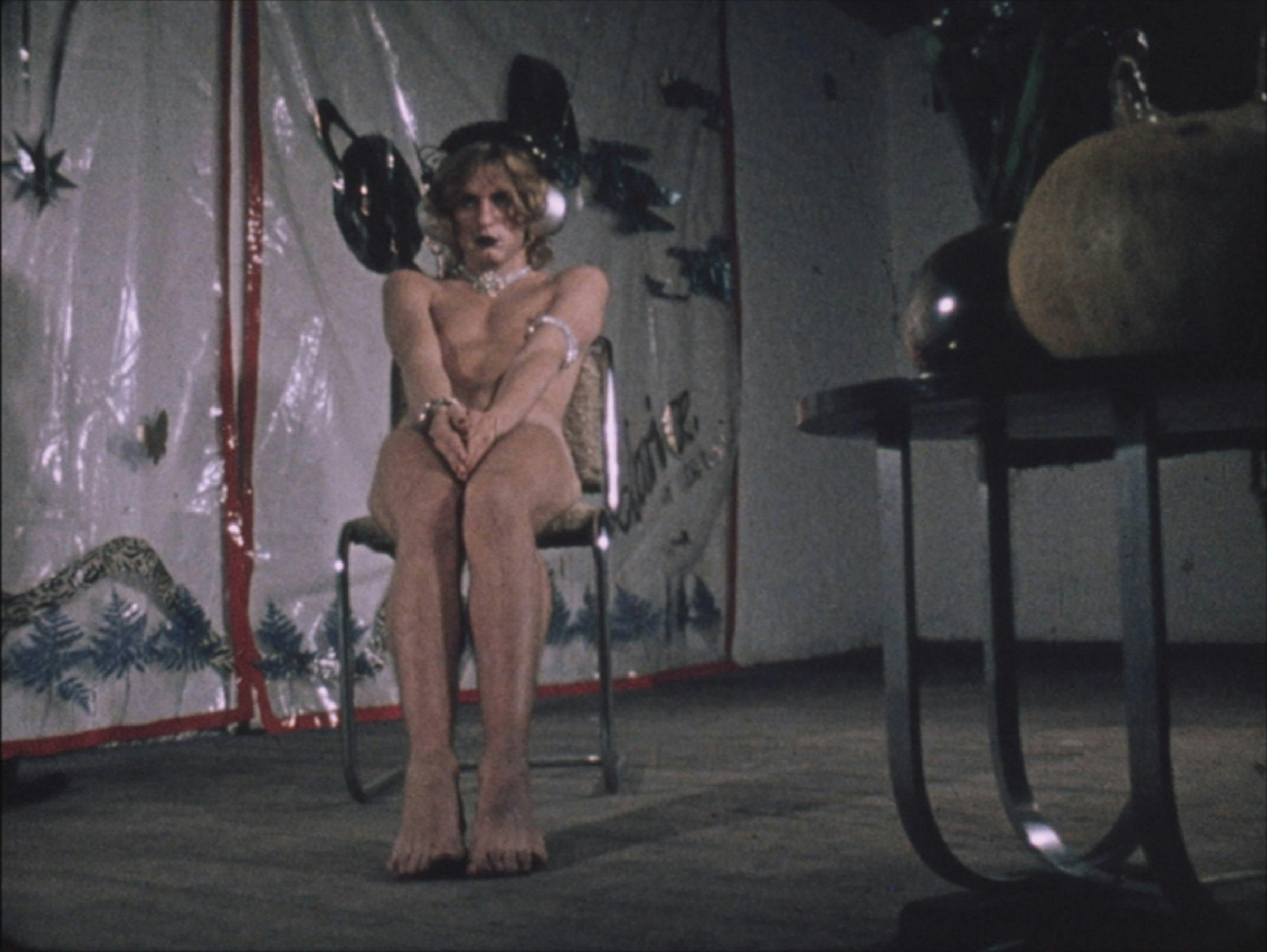 A blond, curly-haired femme person sits naked on a chair. They are sat in a room with a table in the foreground with a vase sat on top of it.