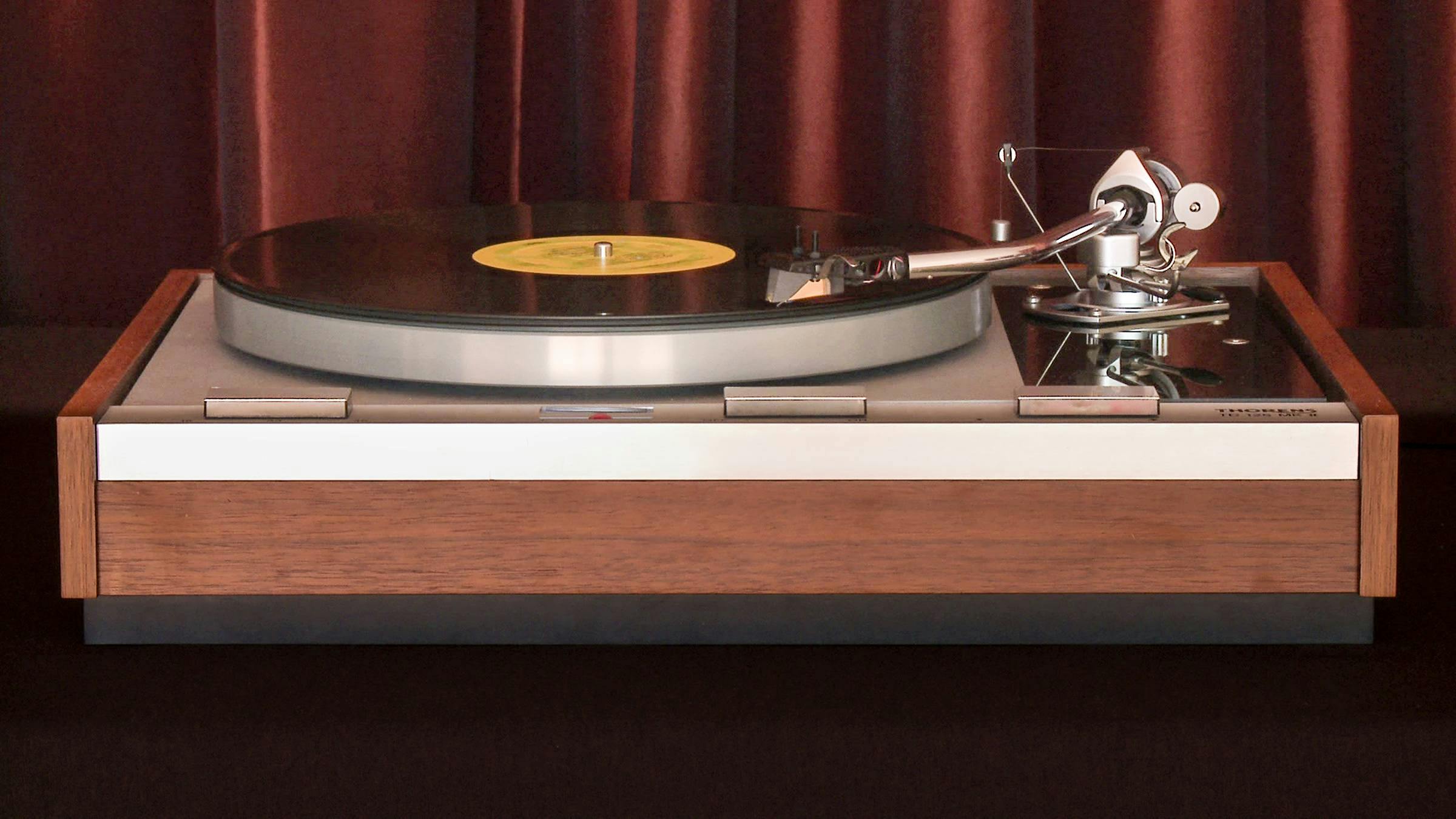a wooden vinyl record player sit on top of a table. There is a red velvet curtain behind it.