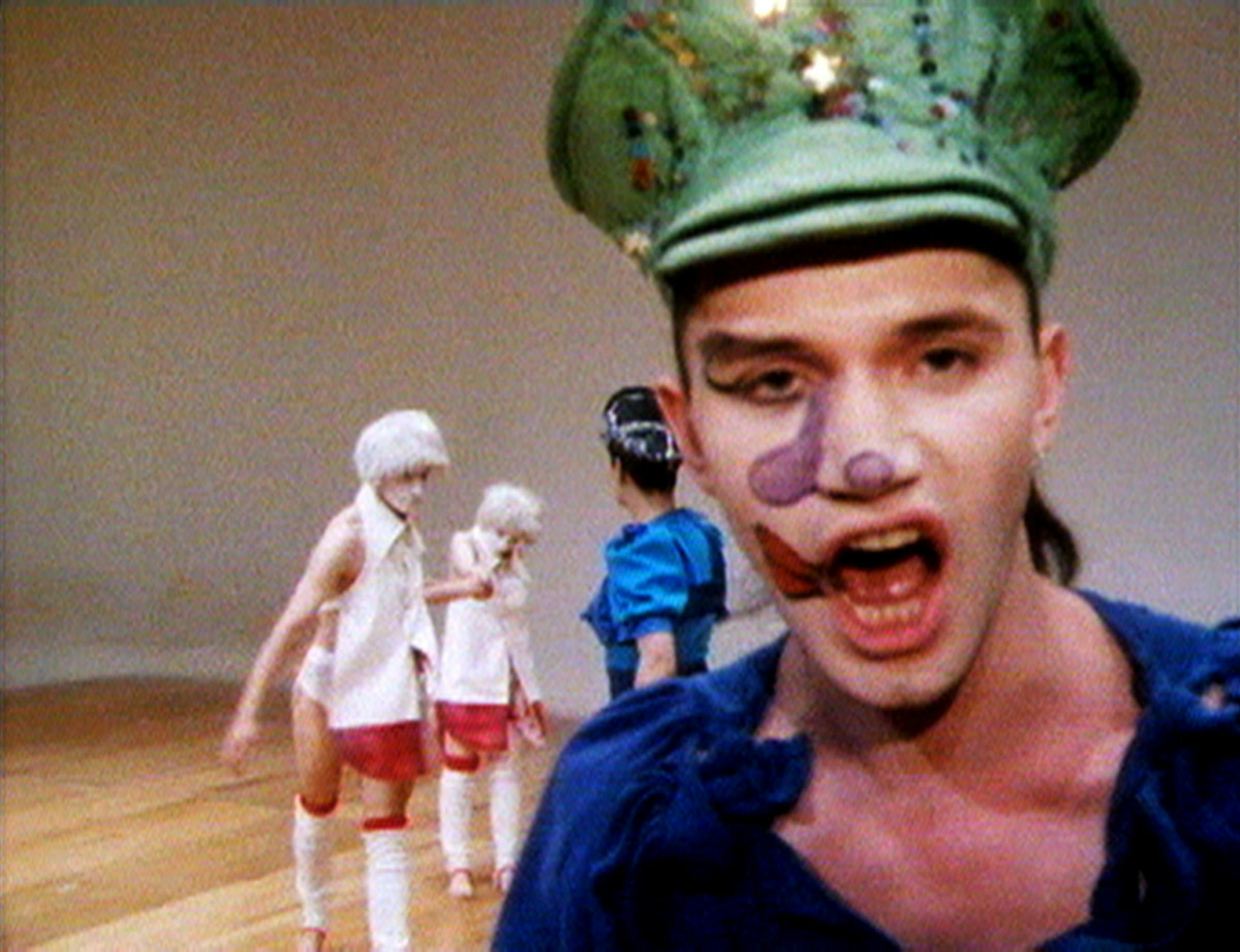 Michael Clark is looking at the camera. He is wearing a green hat and blue top. He is wearing face paint. There are 3 dancers in the background. The floor is wooden.