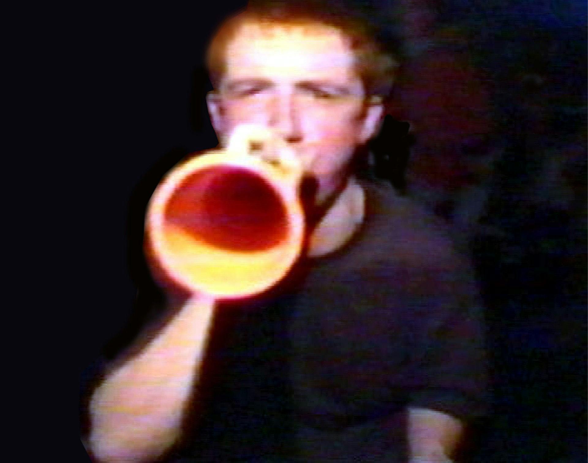 Image of a person with ginger hair and a black t-shirt blowing into a yellow horn whilst looking at the camera.