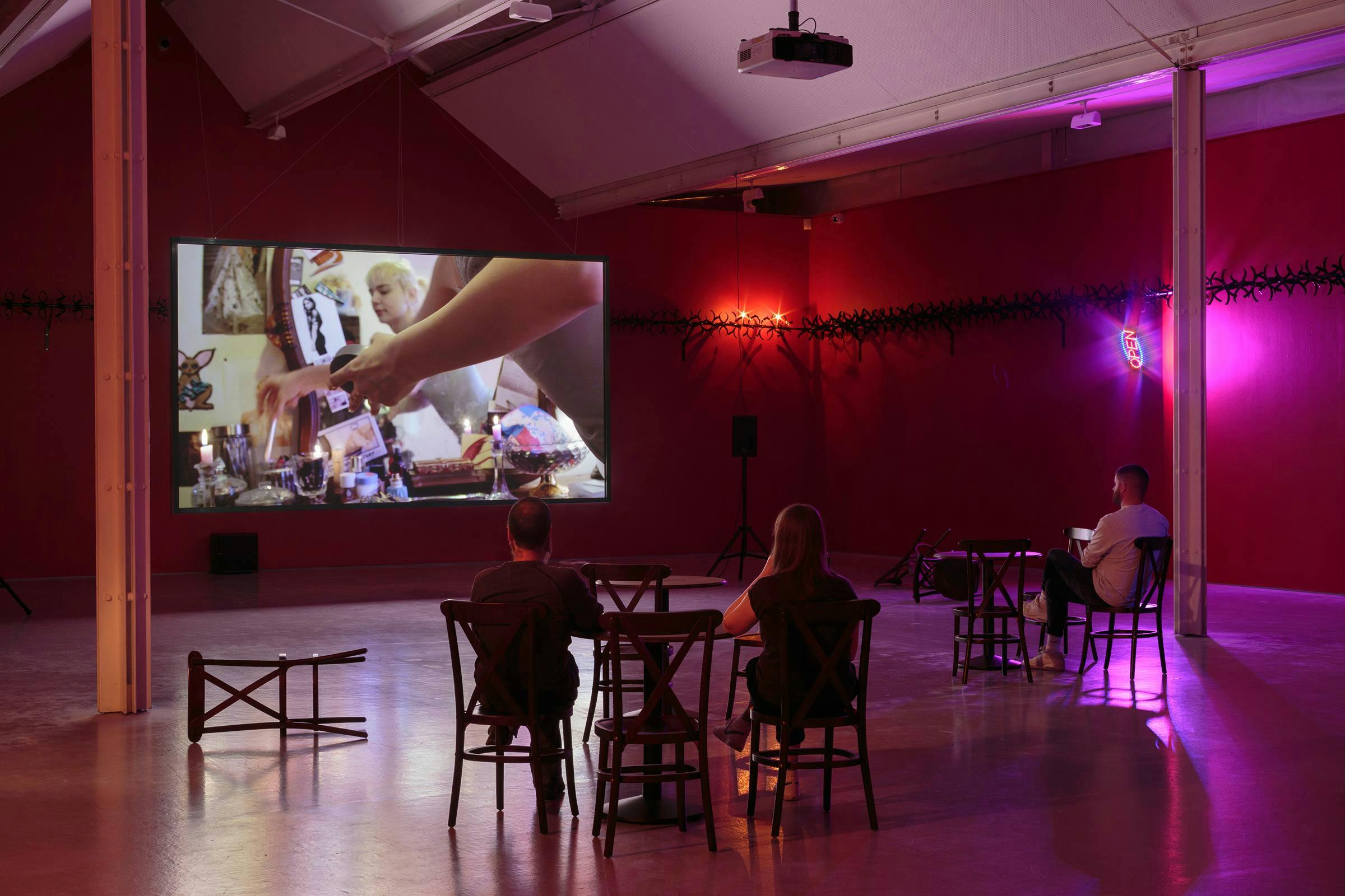 A video art work installed in a gallery space. There are a number of chairs and tables in front of the projection that 3 people are sitting on. The room is tinted with pink light.