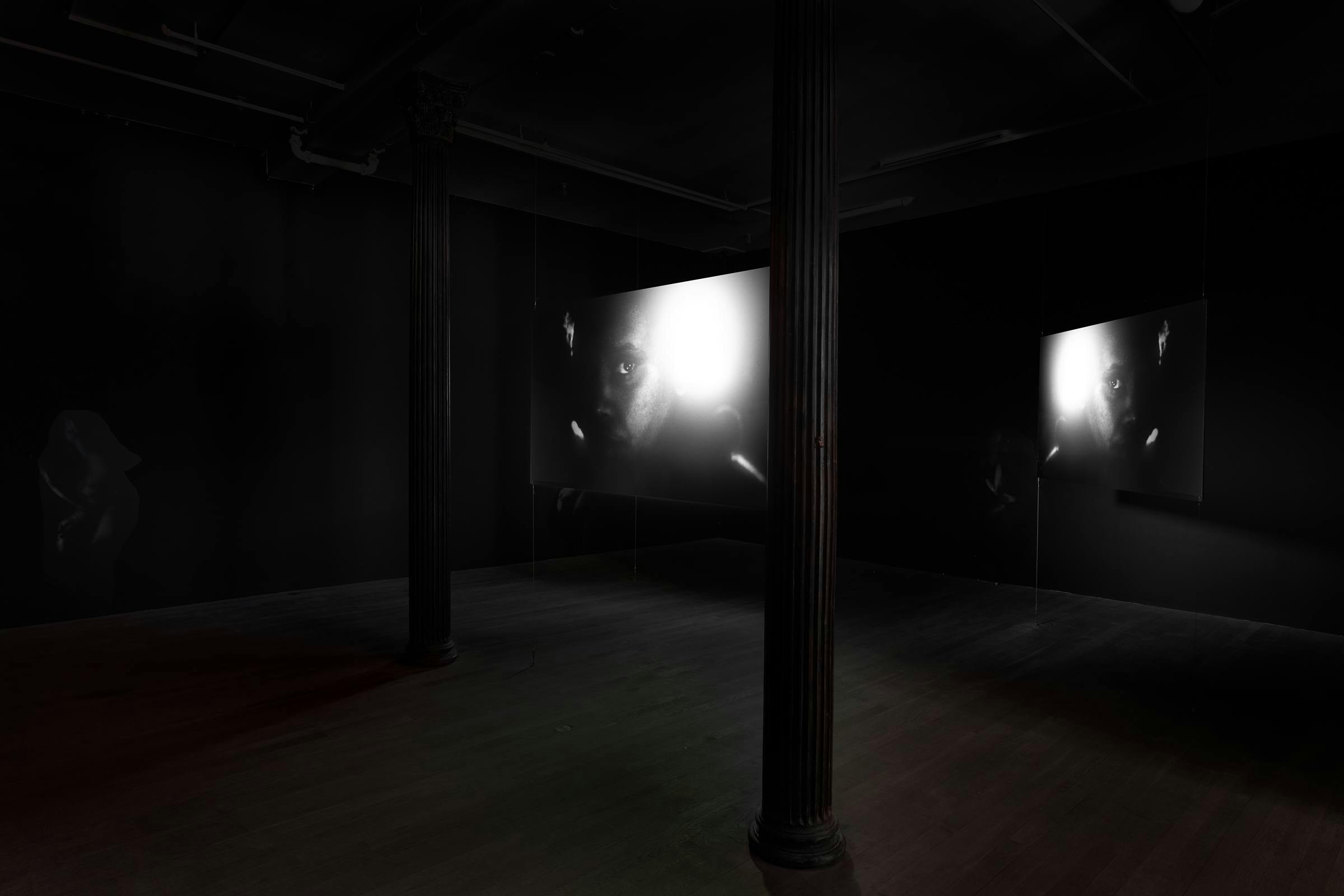Two screens show faces in black and white in a gallery space with columns in it.