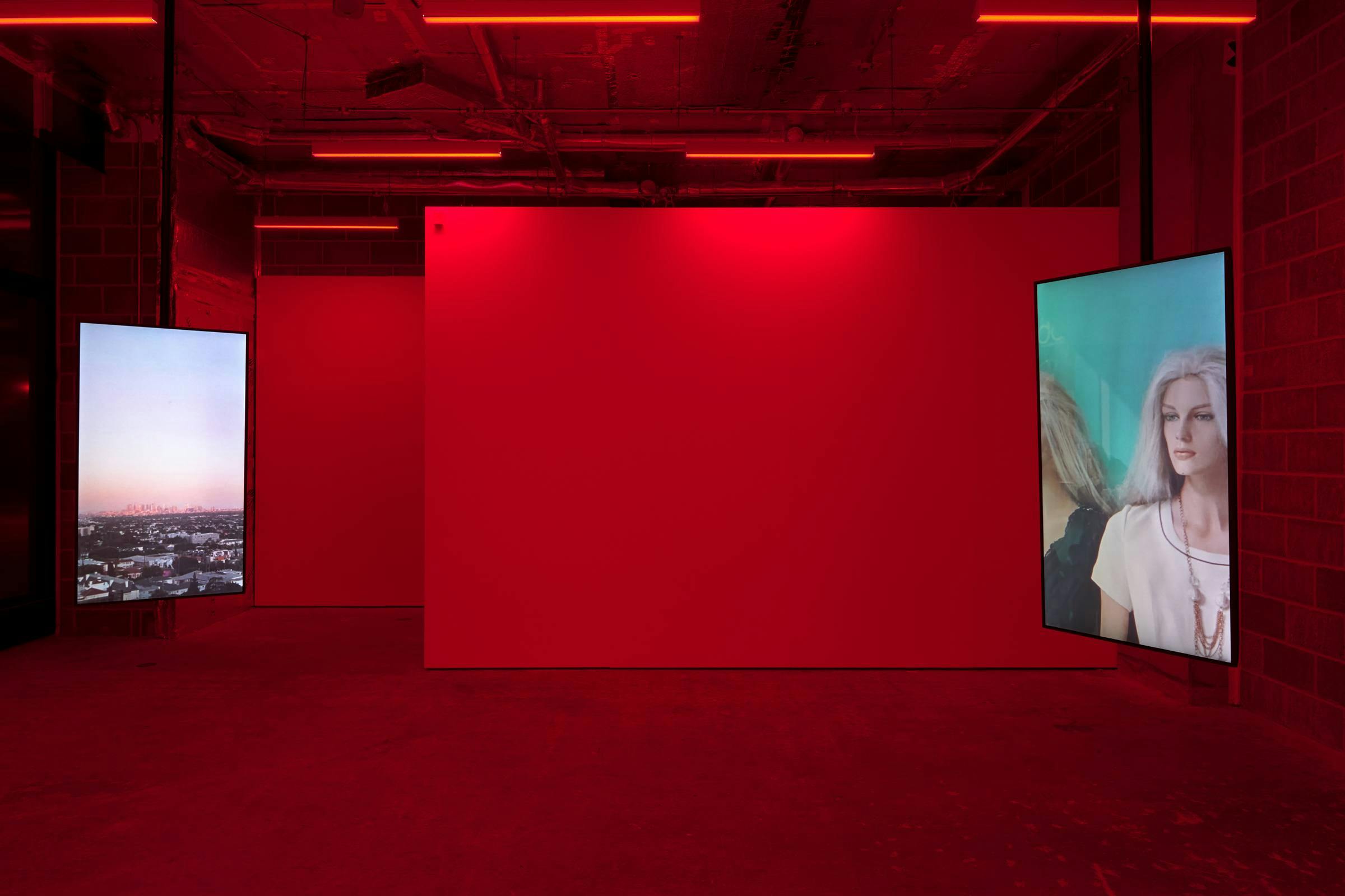 A image of art works in a gallery space. The space is lit red and there are 2 vertical monitors on view that are suspended from the ceiling