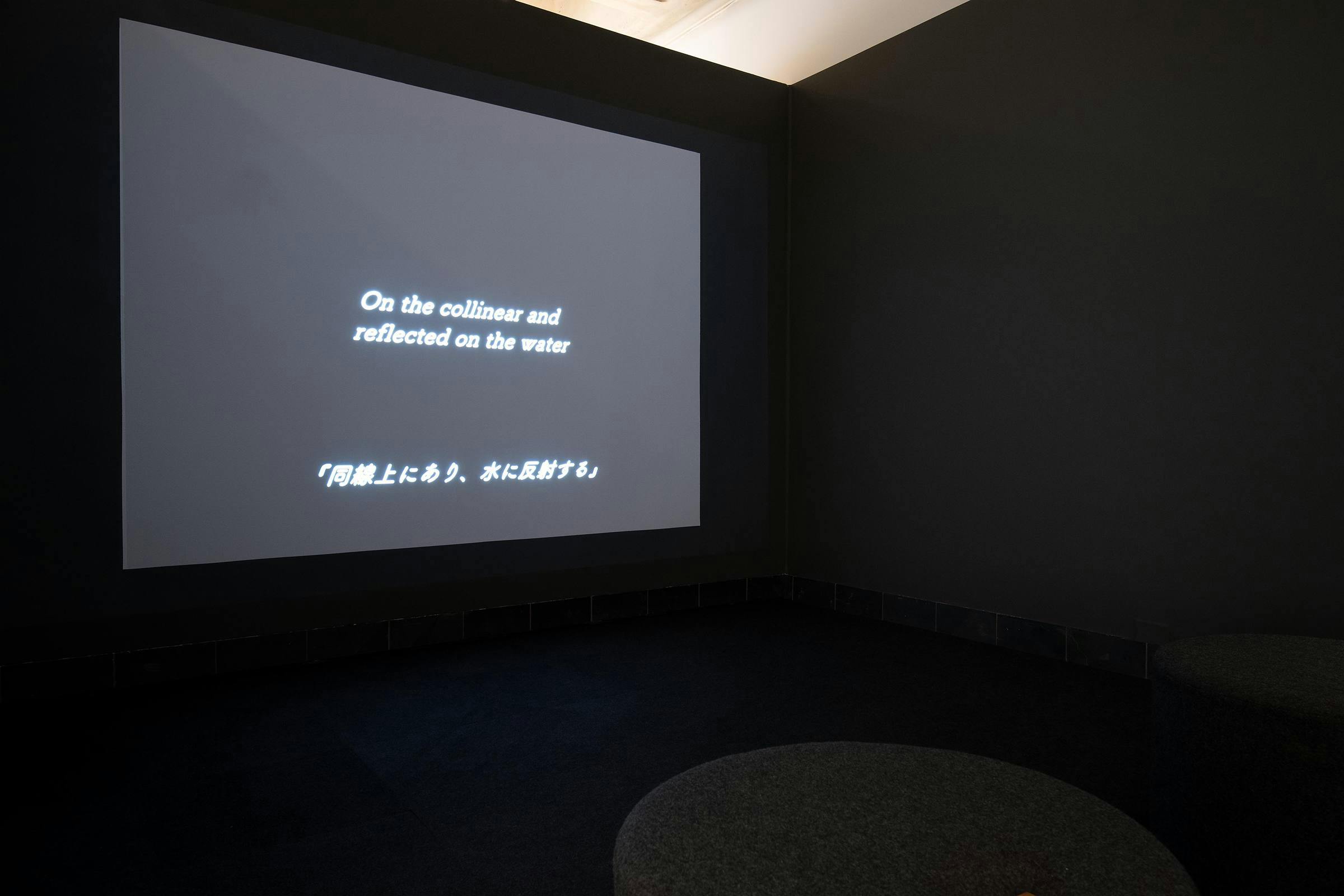 A video artwork projected in a gallery dark space, the projected image is a black screen and a white a subtitle reads 'on the collinear and reflected on the water' there is a Chinese translation underneath the English text