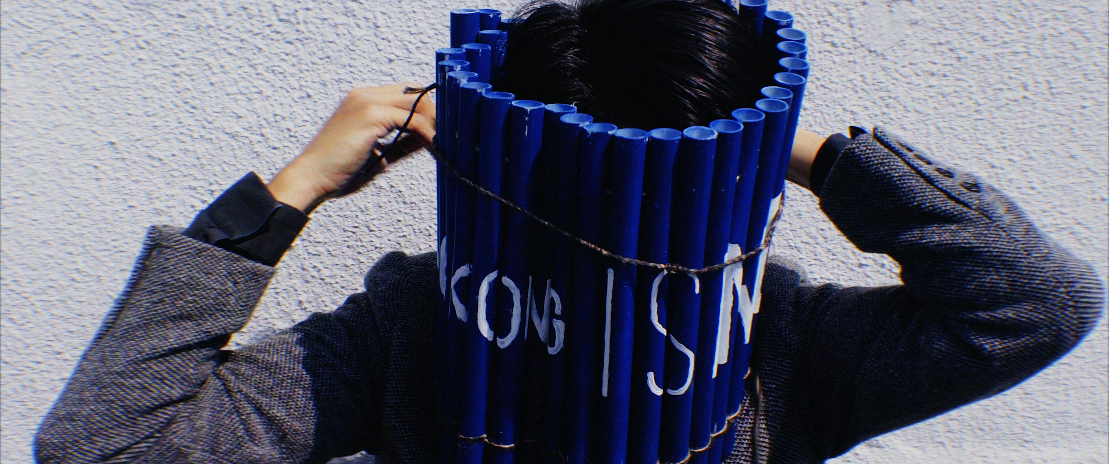 Image of a person tying lots of lengths of blue tubing around their face with some lettering on it. They are wearing a grey blazer.