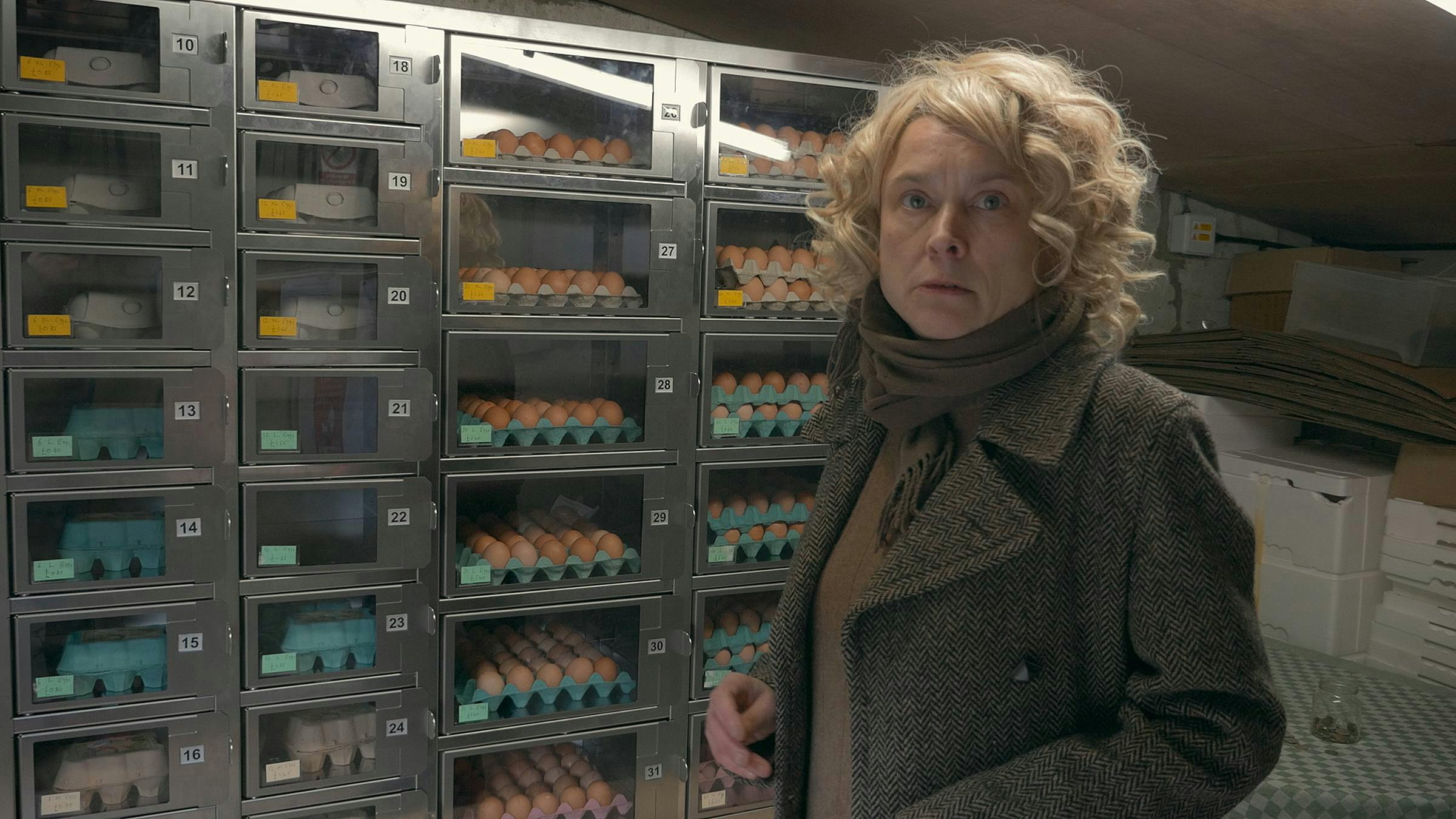 Blond curly haired person stands in the foreground wearing a brown coat. Behind them is a metal case holding eggs in egg trays.